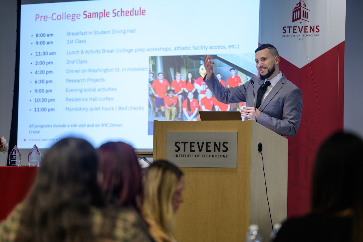 A man speaks at a podium with a screen behind him that reads Pre-College Sample Schedule