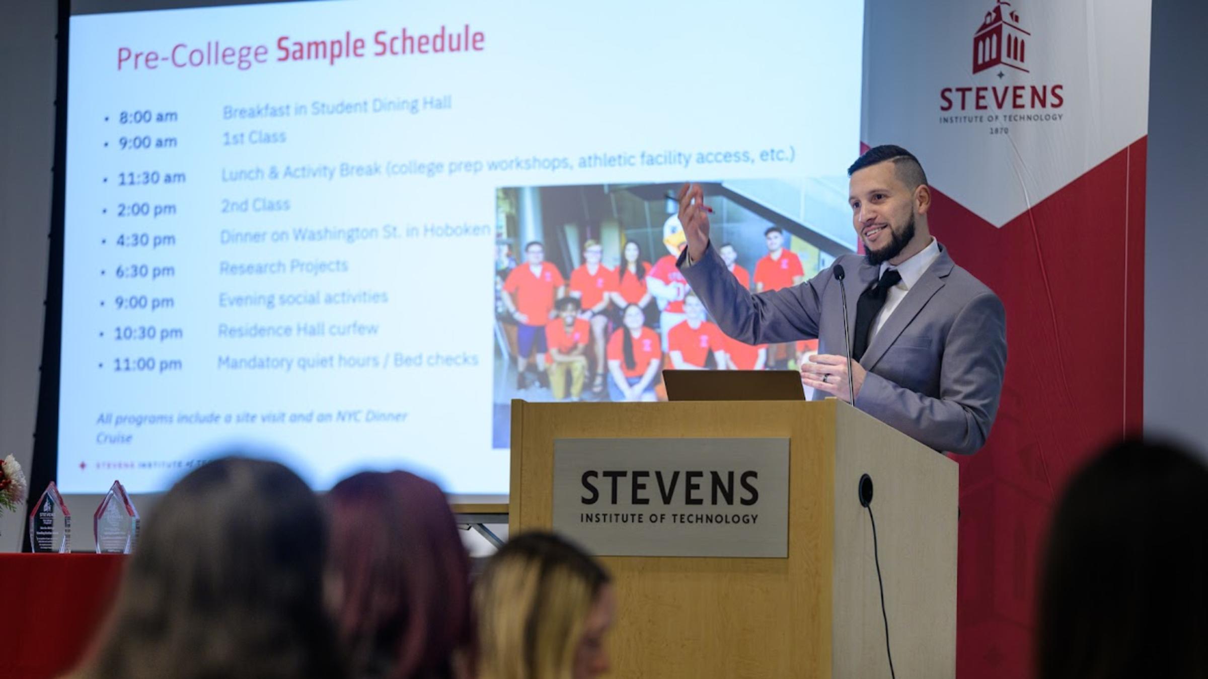 A man speaks at a podium with a screen behind him that reads Pre-College Sample Schedule