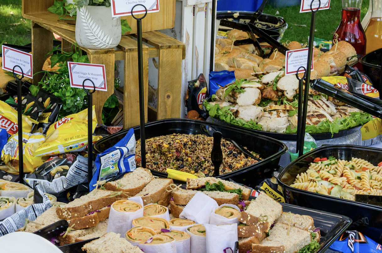 A host of trays containing sandwiches, wraps. grilled chicken, pasta and quinoa at a catered event.