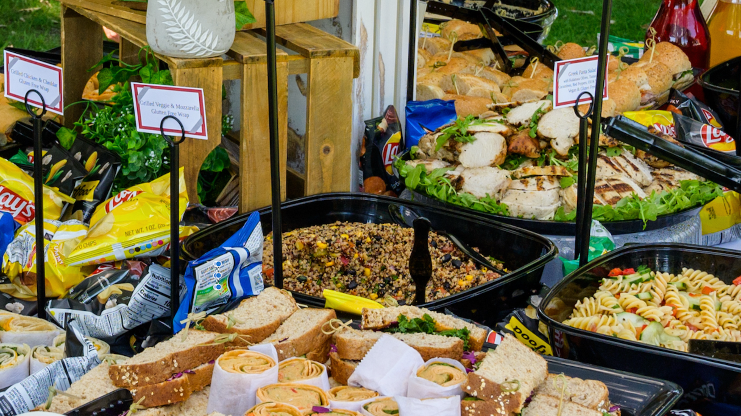 A host of trays containing sandwiches, wraps. grilled chicken, pasta and quinoa at a catered event.