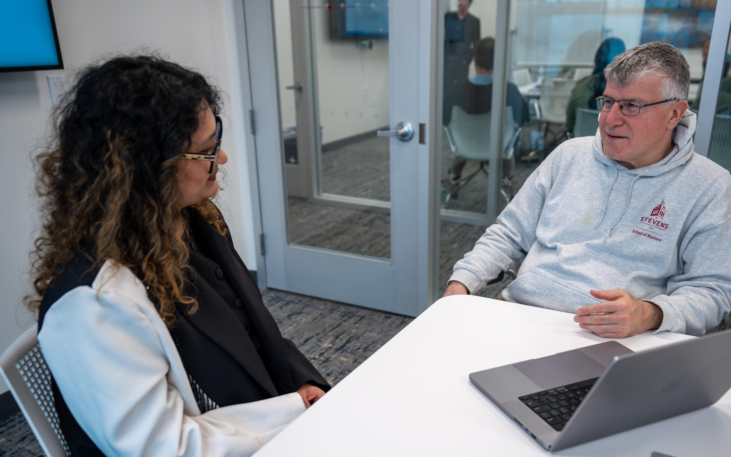 A woman with curly dark hair wearing a dark blazer and white bag sits at a table conversing with a man in a gray Stevens School of Business hoodie and glasses. A laptop is visible on the white table between them in a modern office setting.
