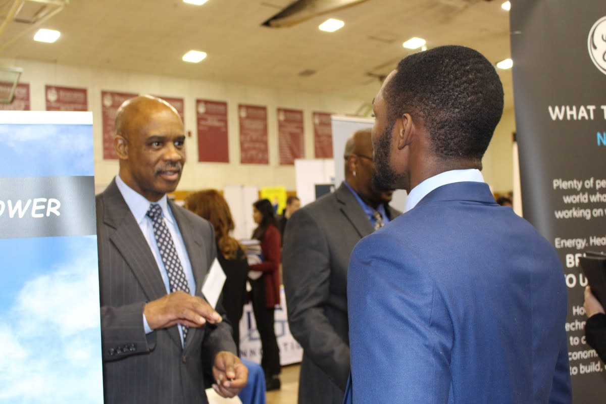 A male recruiter in a gray pinstriped suit, who is standing on the right, speaks to a male student wearing a blue suit, who is facing him across the table