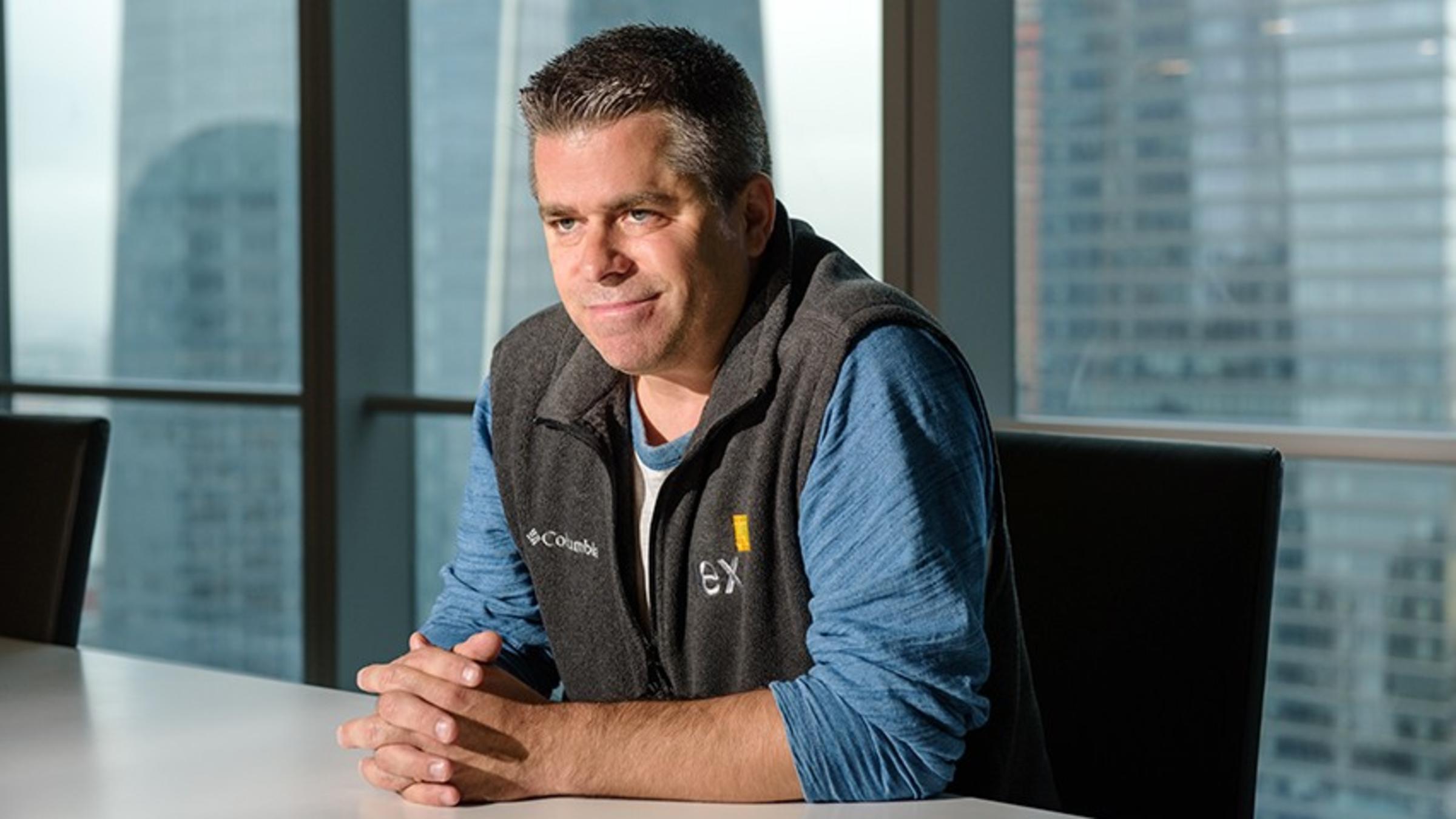 John Schwall in a gray IEX vest and blue shirt at a conference table with the New York City skyline seen through a window.