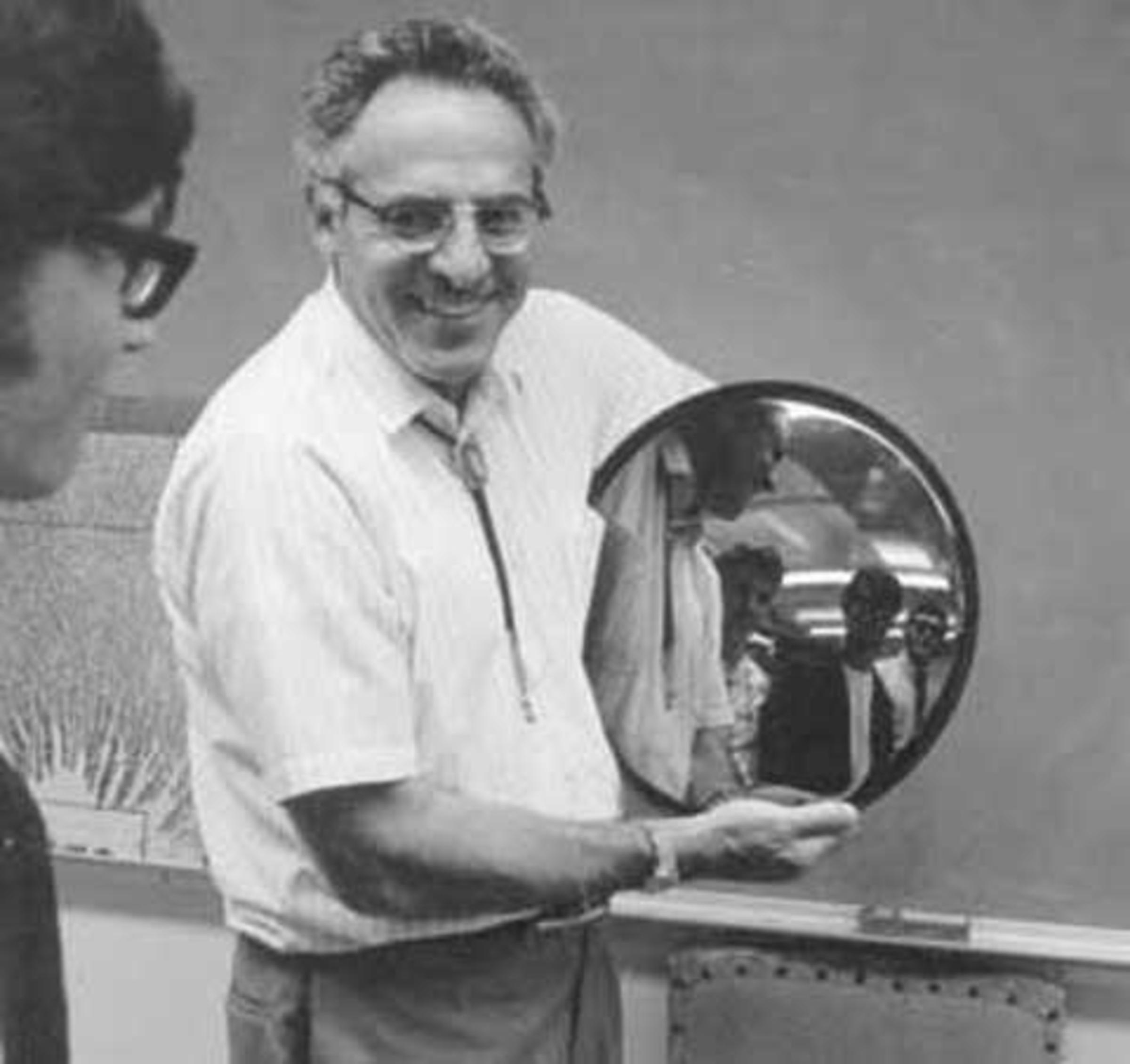 A black-and-white photo of Frederick Reines smiling and holding a mirror that is reflecting the faces of people standing in front of him.