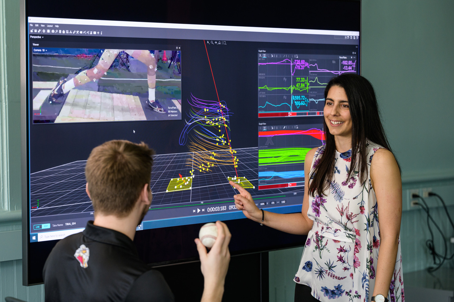 Antonia Zaferiou in her laboratory pointing to a screen with data models as a student holds a baseball in the foreground