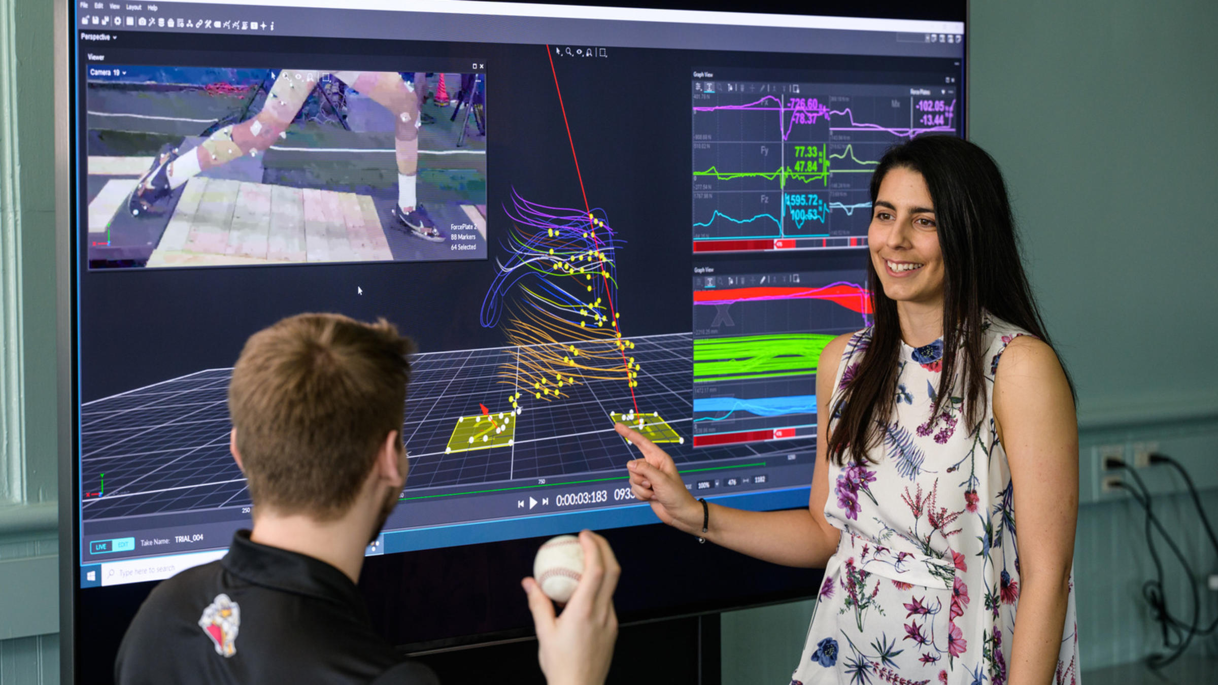 Antonia Zaferiou in her laboratory pointing to a screen with data models as a student holds a baseball in the foreground
