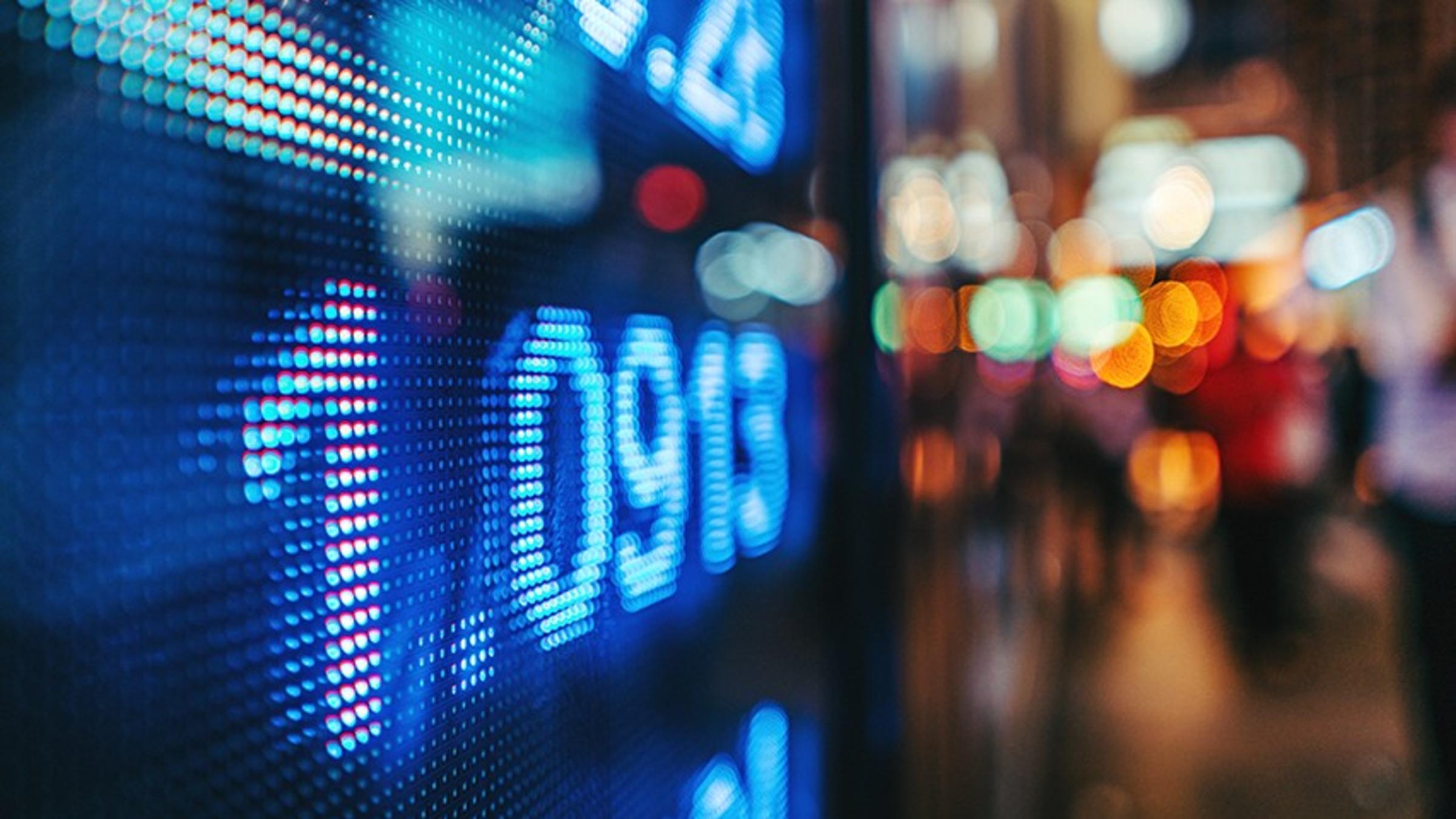 Digital stock ticker displayed from a screen on a darkened street.