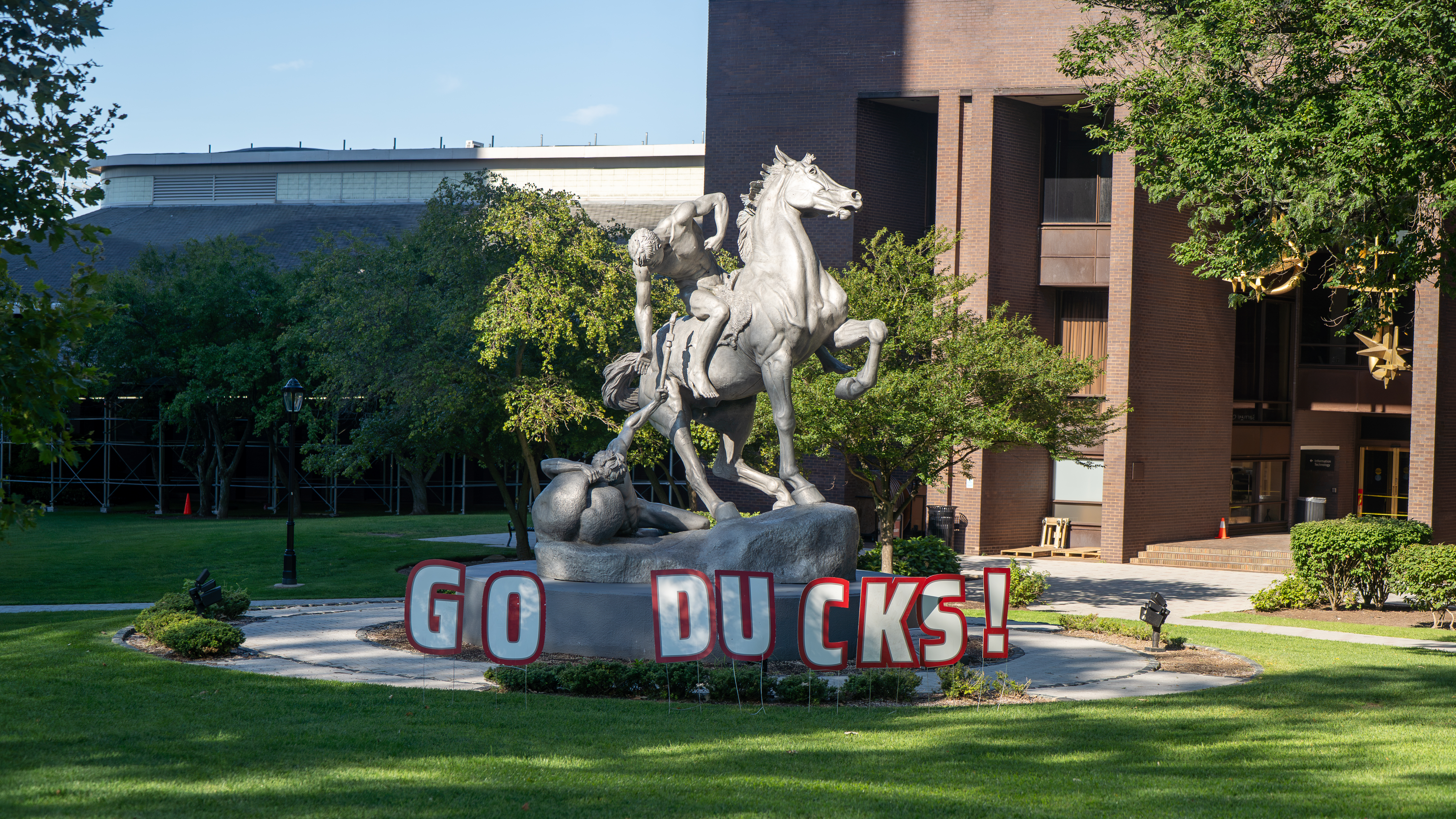 A silver statue of a rearing horse with two figures in a struggle, on a stone pedestal with "GO DUCKS!" letters in front, set on a lawn near a brick campus building with trees.