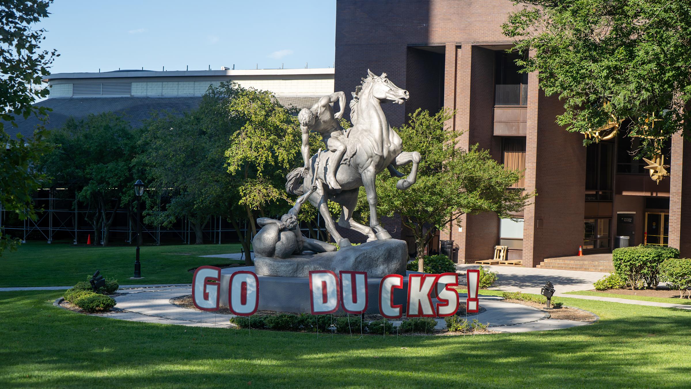 A silver statue of a rearing horse with two figures in a struggle, on a stone pedestal with "GO DUCKS!" letters in front, set on a lawn near a brick campus building with trees.