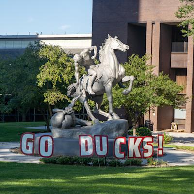 A silver statue of a rearing horse with two figures in a struggle, on a stone pedestal with "GO DUCKS!" letters in front, set on a lawn near a brick campus building with trees.