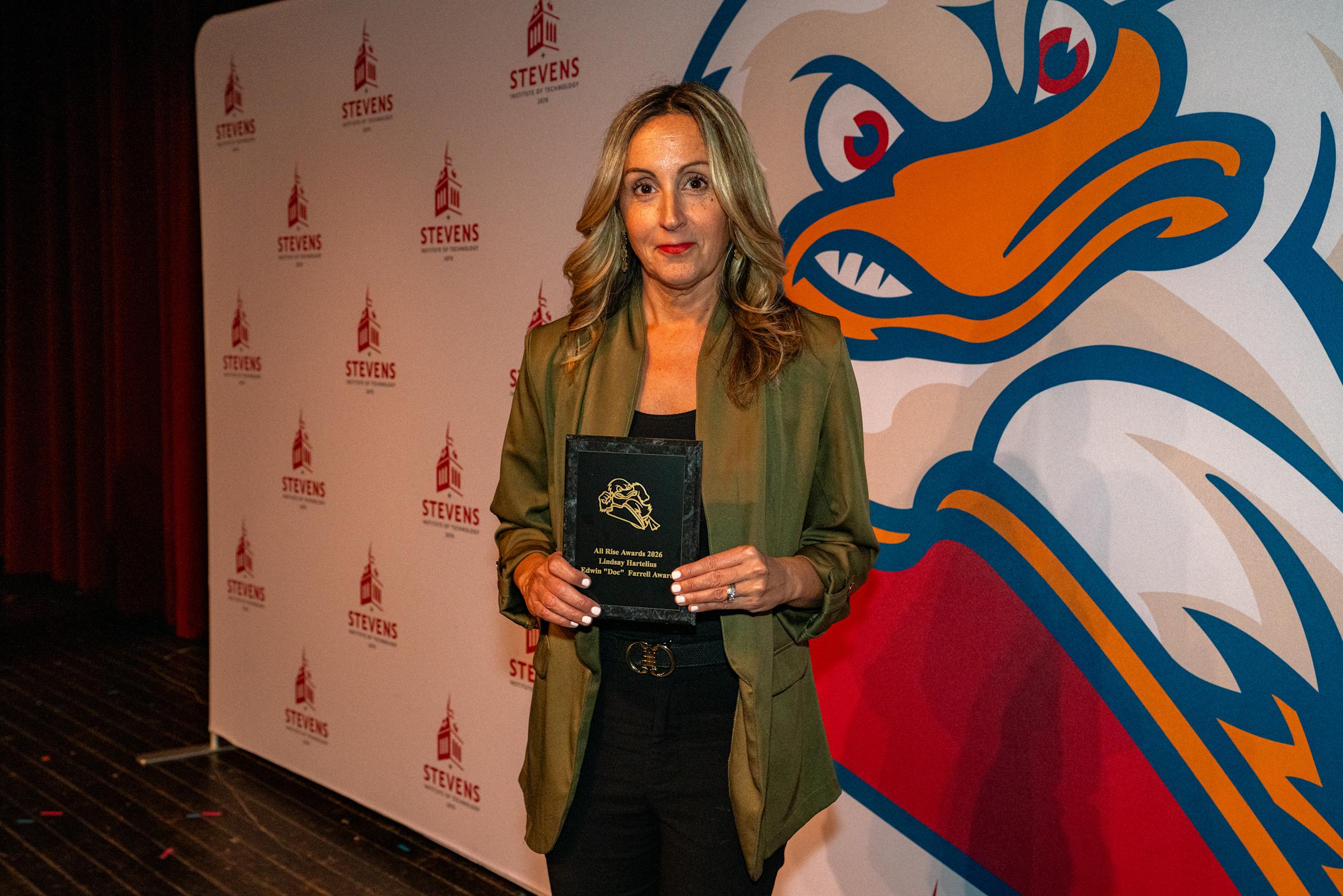 Woman in olive green blazer over black outfit holds Stevens Athletics All Rise Awards 2026 black plaque with gold design, standing in front of step-and-repeat backdrop featuring Stevens Institute logos and duck mascot graphic in blue, orange, and red.