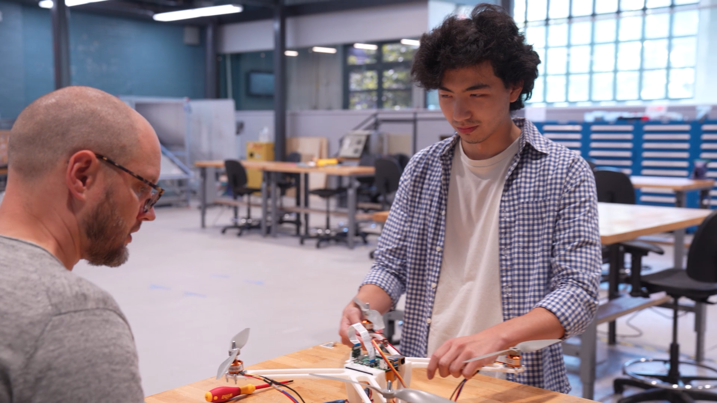 A young man in a checkered shirt demonstrates a drone or electronic device project to a bald instructor with glasses in a makerspace, with workbenches, tool cabinets, and large windows visible in the bright, industrial workspace.