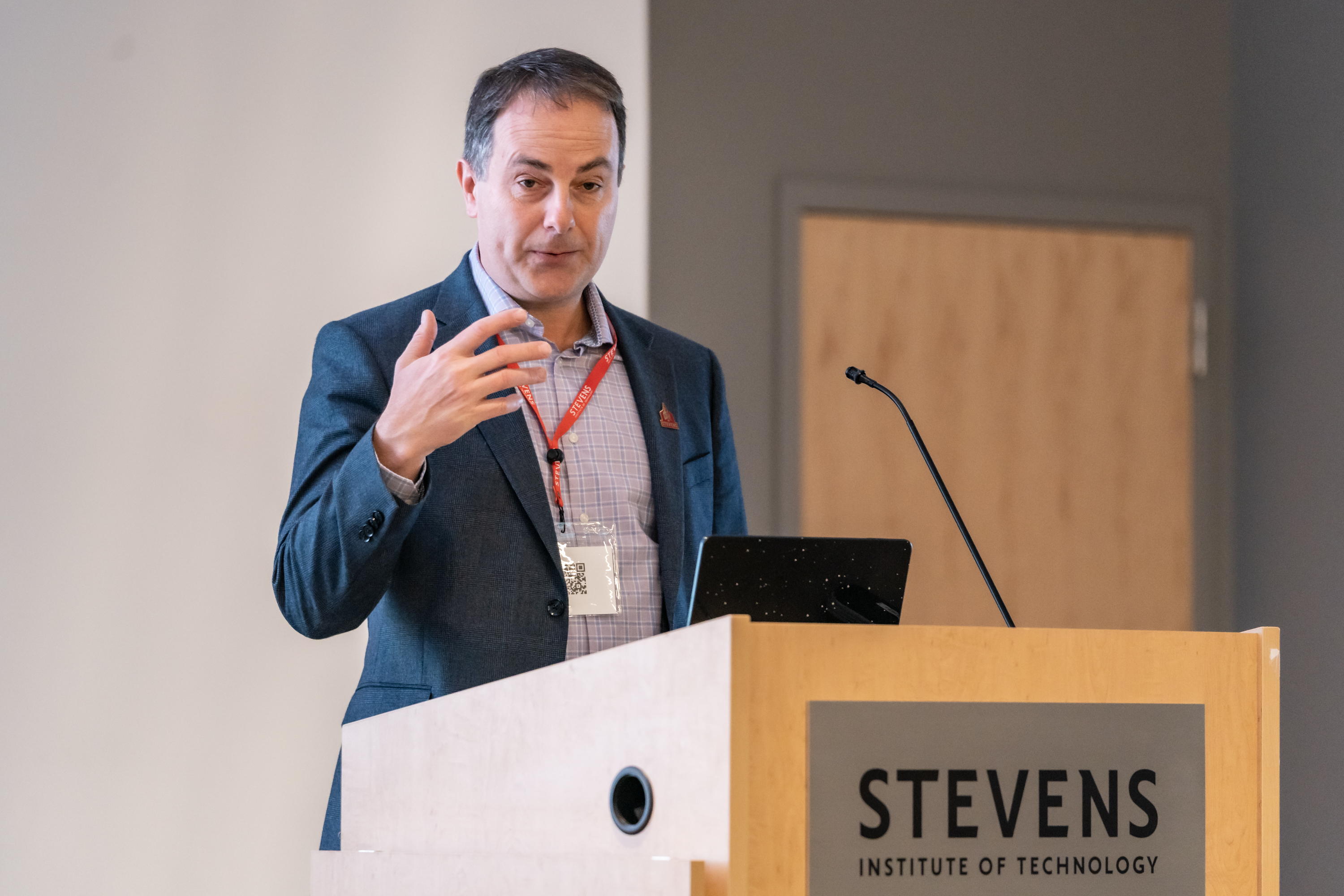 A man in a dark suit jacket and checkered shirt wearing a red conference lanyard presents at a wooden podium bearing the Stevens Institute of Technology logo. He gestures while speaking, with a laptop and microphone on the podium before him.