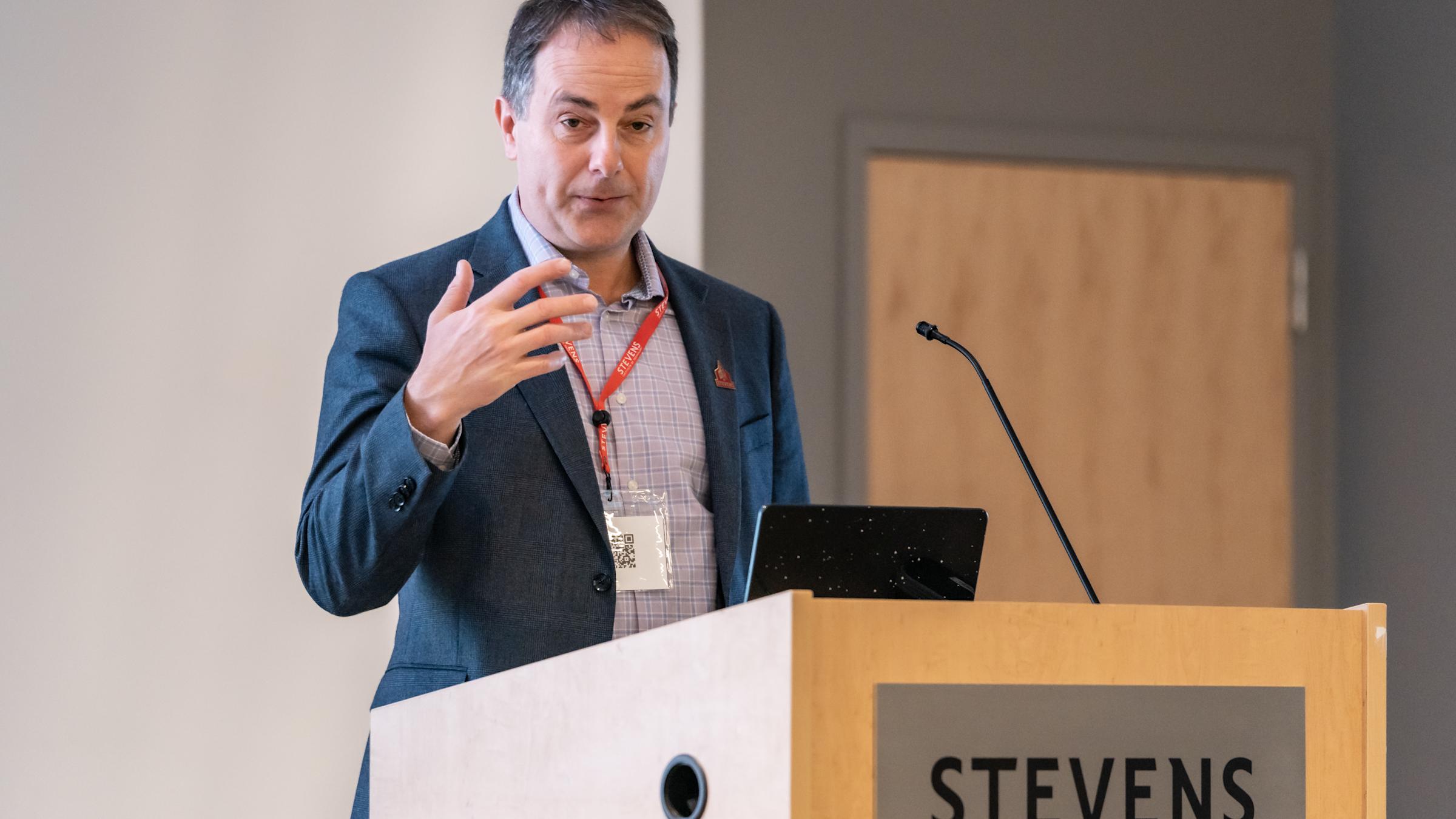 A man in a dark suit jacket and checkered shirt wearing a red conference lanyard presents at a wooden podium bearing the Stevens Institute of Technology logo. He gestures while speaking, with a laptop and microphone on the podium before him.