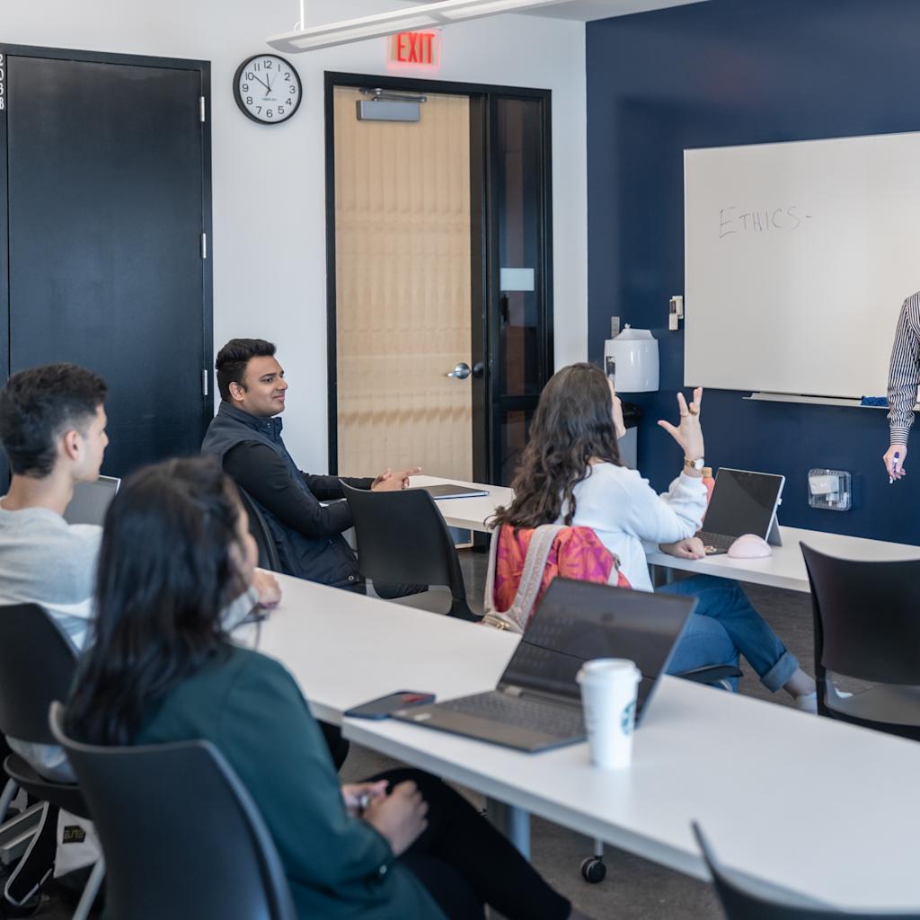 A man in a striped shirt presents to a group of students or professionals seated at tables with laptops in a modern classroom. One participant raises their hand while others listen attentively. A whiteboard shows "Ethics" written on it.