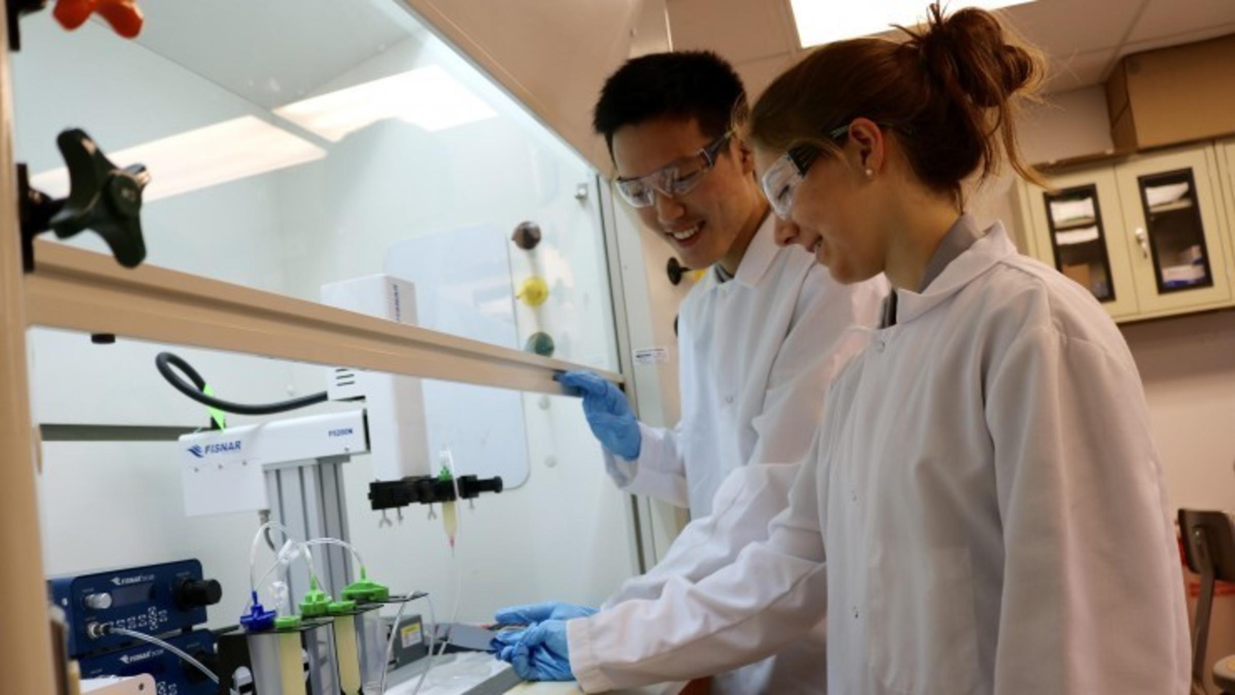 A photo of two students wearing lab gear looking at test tubes