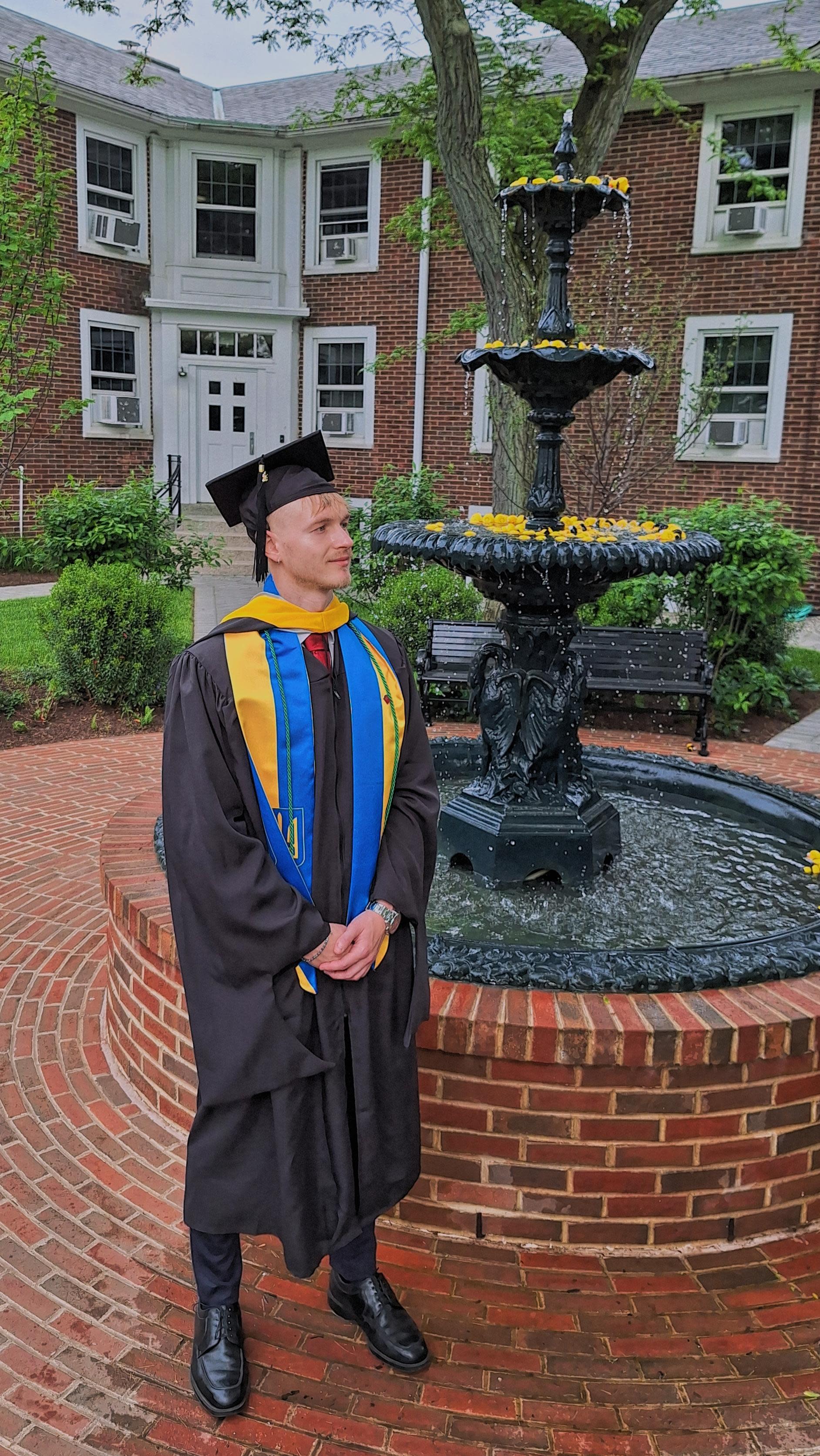 "Graduate in black cap and gown with blue and gold academic hood stands beside ornate tiered fountain on brick campus courtyard with red brick buildings in background"