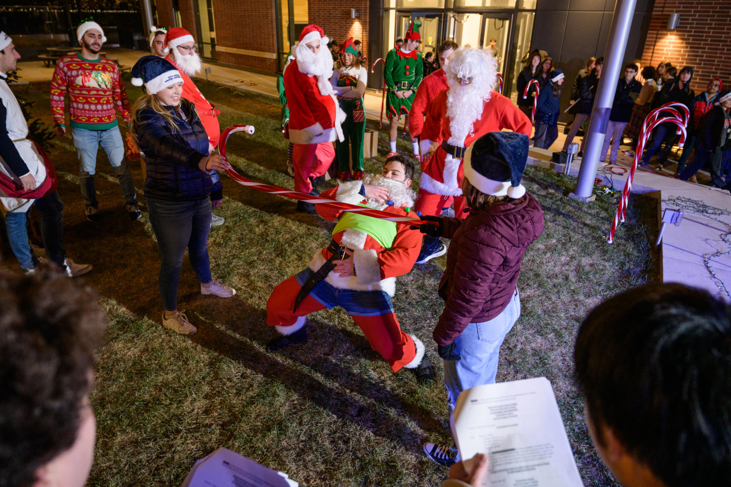 A student Santa competition in progress, with participants dressed in Santa costumes competing in a limbo contest. One student is leaning back to pass under a horizontal bar while others cheer on from the sidelines.