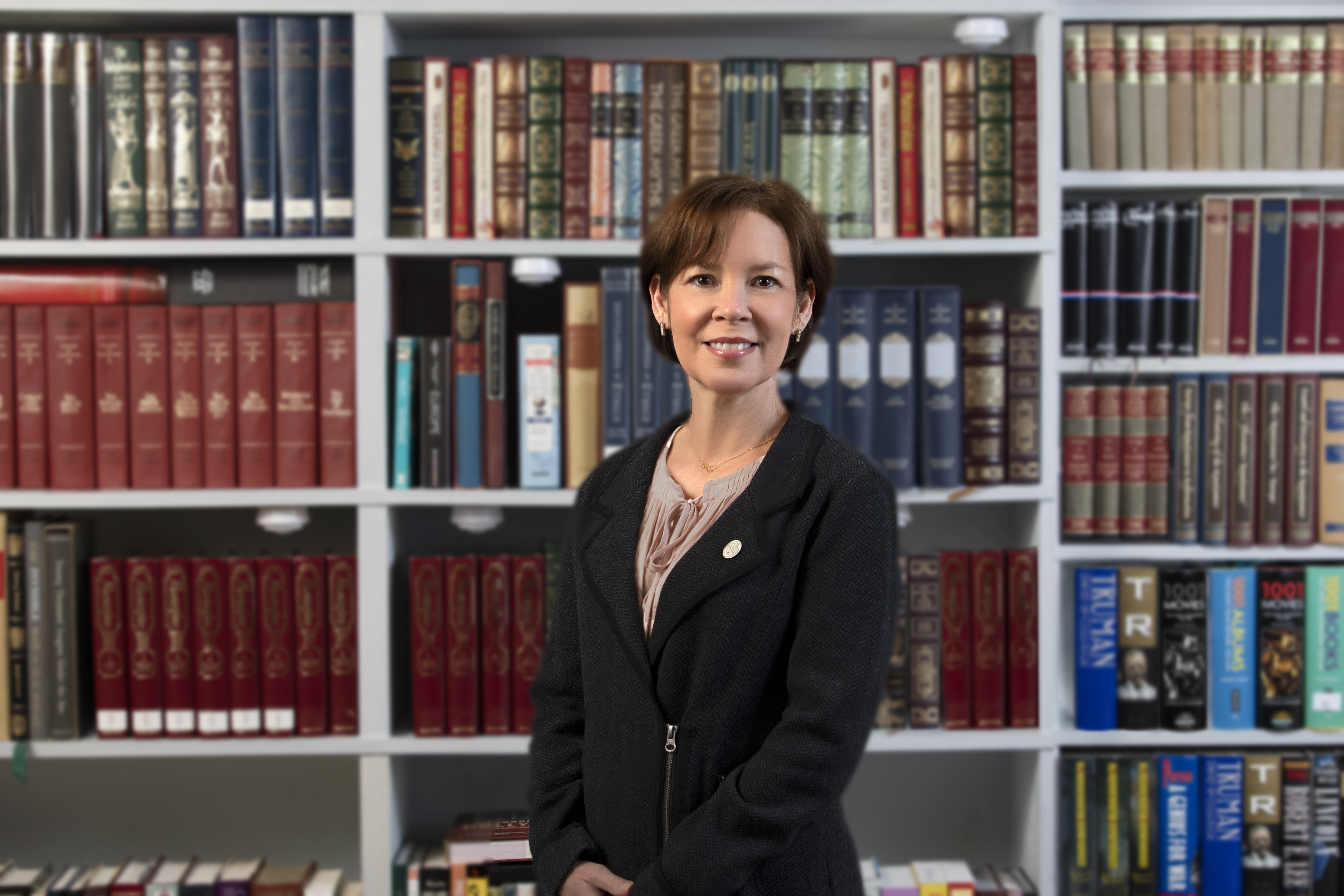 A woman, Beth McGrath, posing in front of a bookcase filled with books. She is wearing a black jacket.