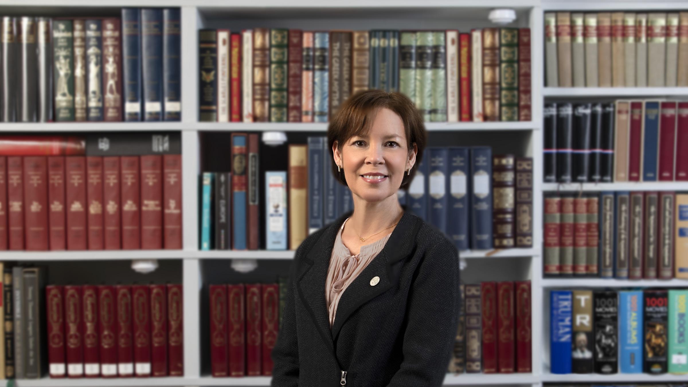 A woman, Beth McGrath, posing in front of a bookcase filled with books. She is wearing a black jacket.