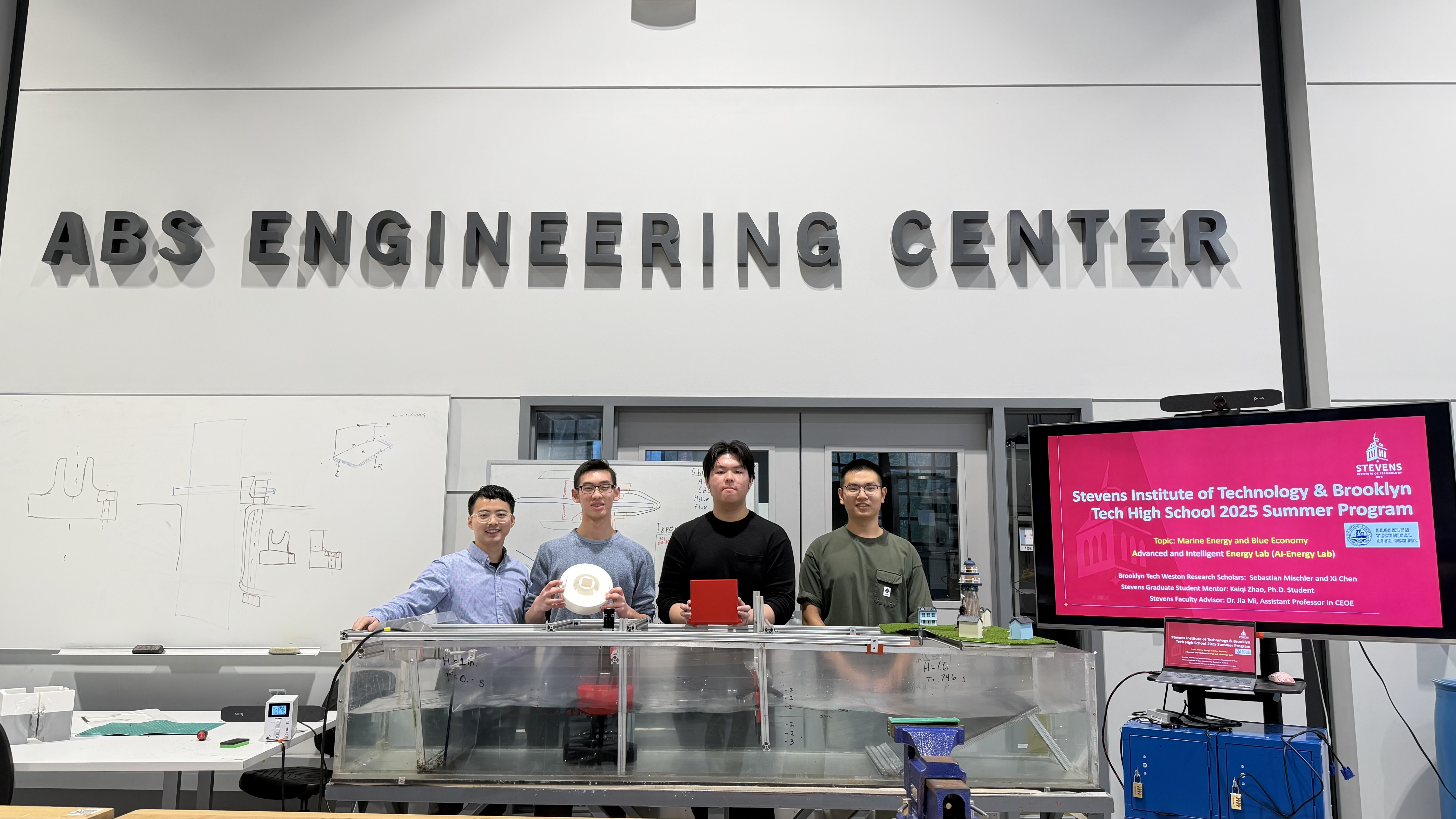 Jia Mi and a group of Brooklyn Tech High School students stand together in the ABS Engineering Center next to a presentation monitor.