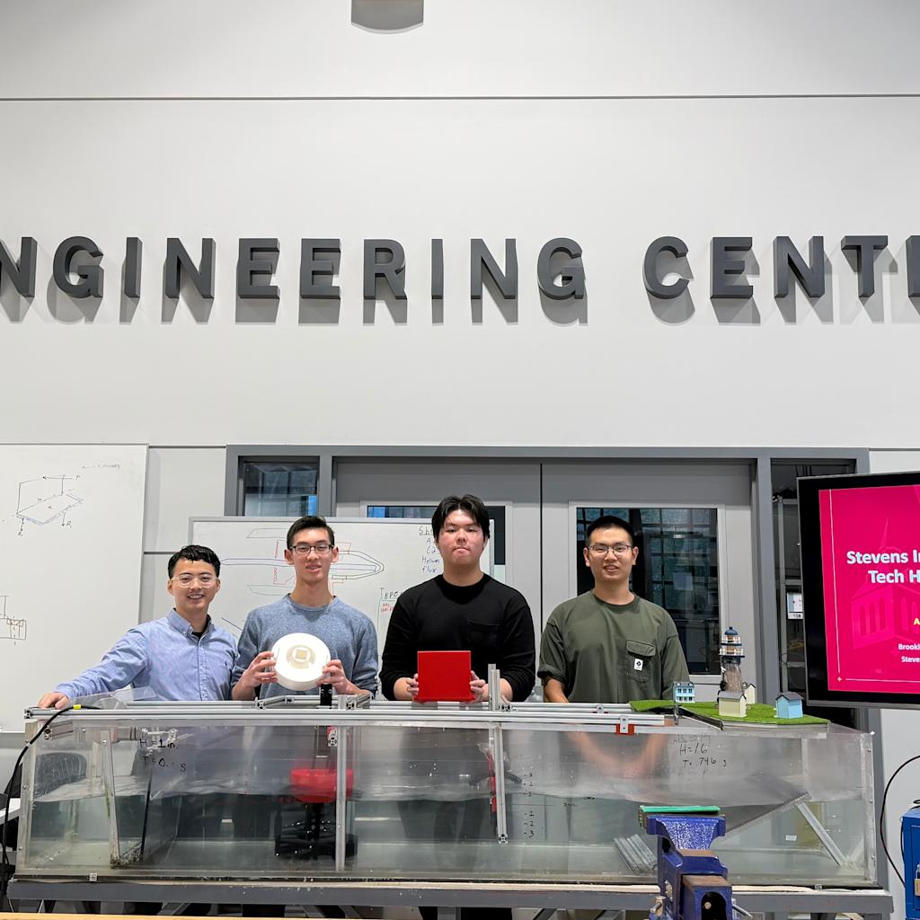 Jia Mi and a group of Brooklyn Tech High School students stand together in the ABS Engineering Center next to a presentation monitor.