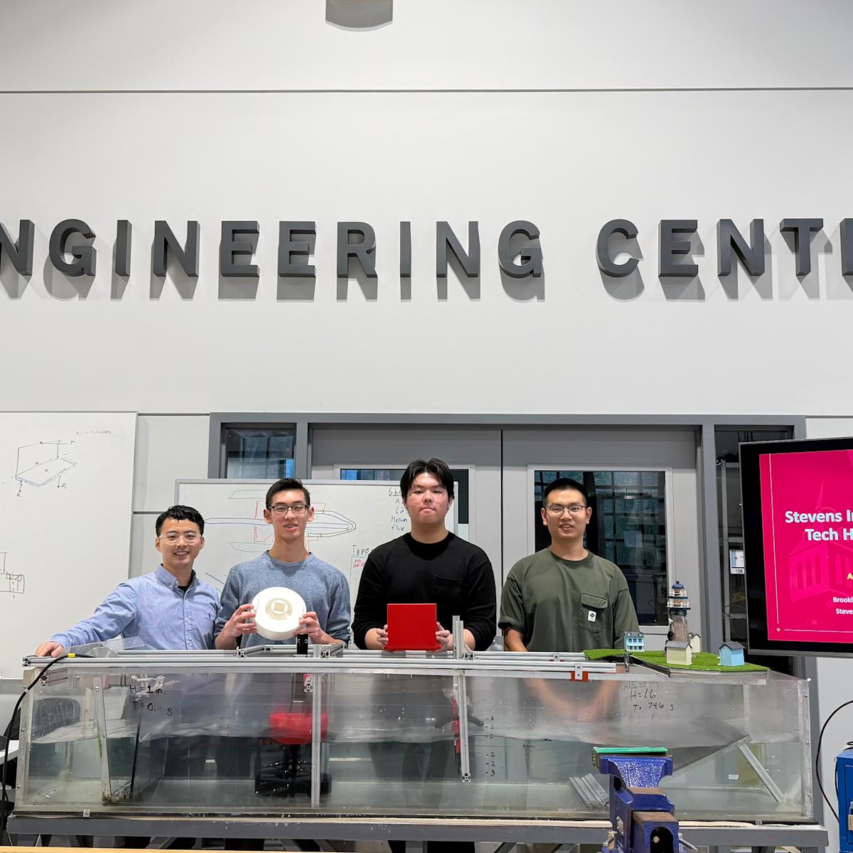 Jia Mi and a group of Brooklyn Tech High School students stand together in the ABS Engineering Center next to a presentation monitor.