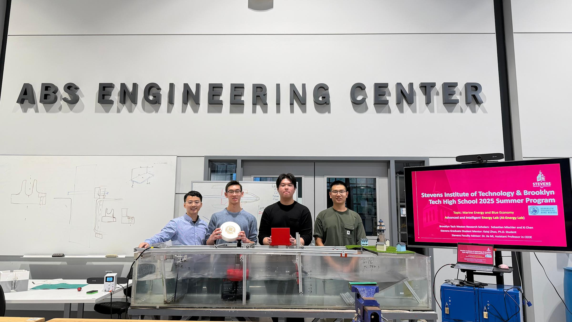 Jia Mi and a group of Brooklyn Tech High School students stand together in the ABS Engineering Center next to a presentation monitor.