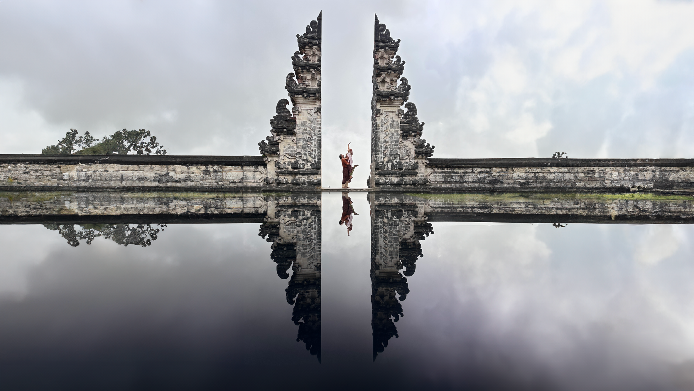 A man lifts a woman in the air in the distance, as they stand near an ornate wall, with a body of water in front of them showing their reflection.