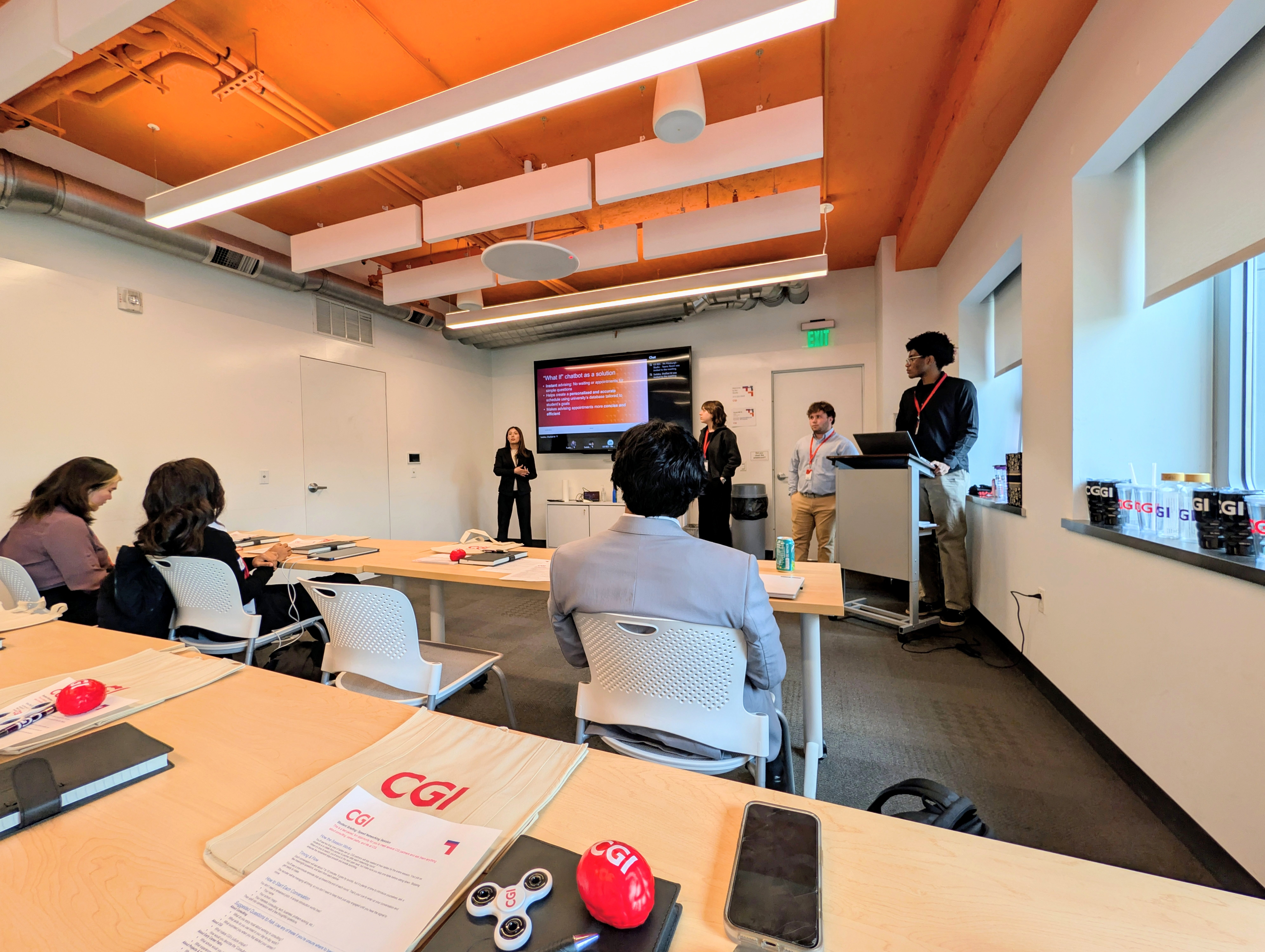 Students present to a small audience in a modern conference room with exposed wood beam ceiling and accent lighting. CGI-branded materials including folders, stress balls, and water bottles are arranged on wood tables. A presentation slide is displayed.