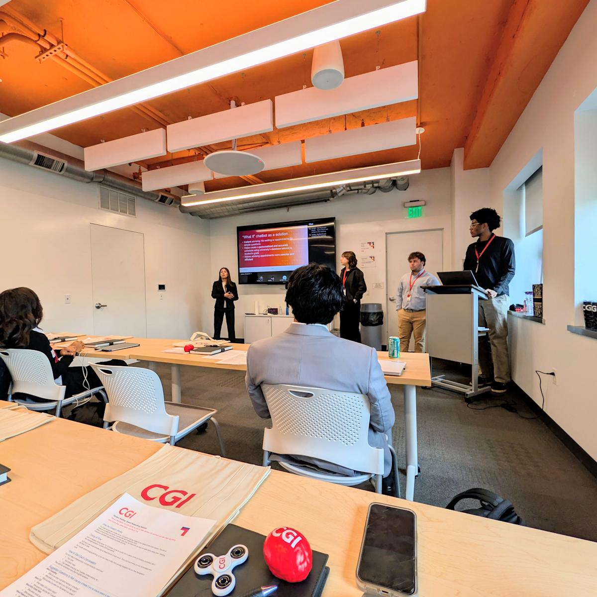 Students present to a small audience in a modern conference room with exposed wood beam ceiling and accent lighting. CGI-branded materials including folders, stress balls, and water bottles are arranged on wood tables. A presentation slide is displayed.