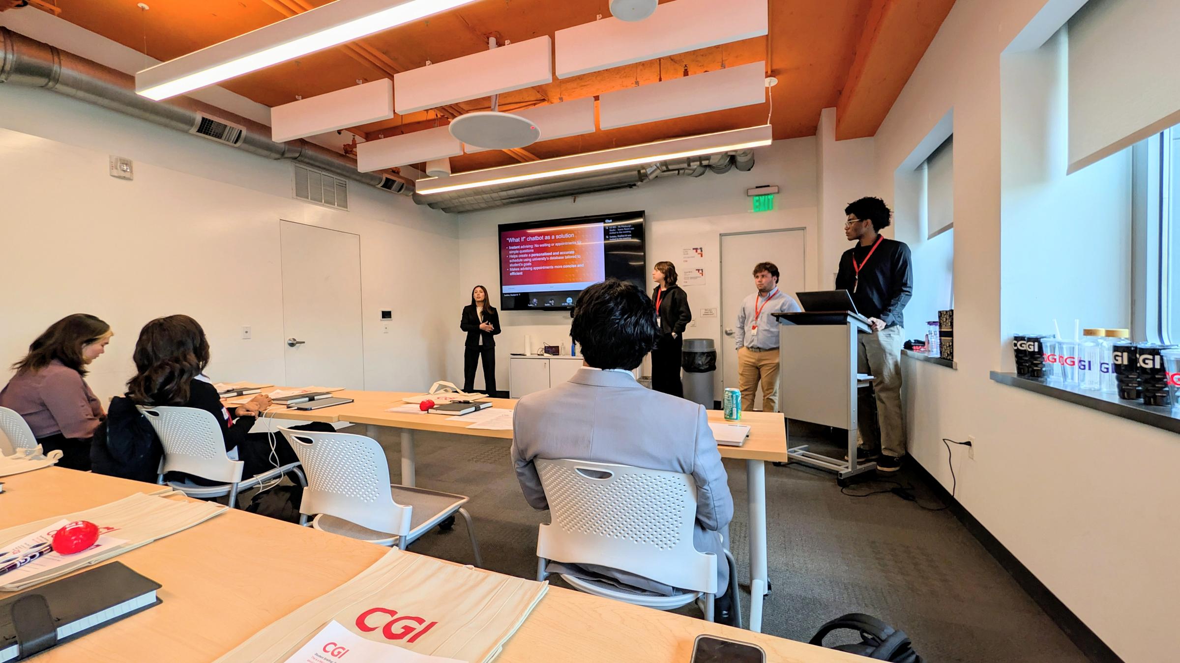 Students present to a small audience in a modern conference room with exposed wood beam ceiling and accent lighting. CGI-branded materials including folders, stress balls, and water bottles are arranged on wood tables. A presentation slide is displayed.