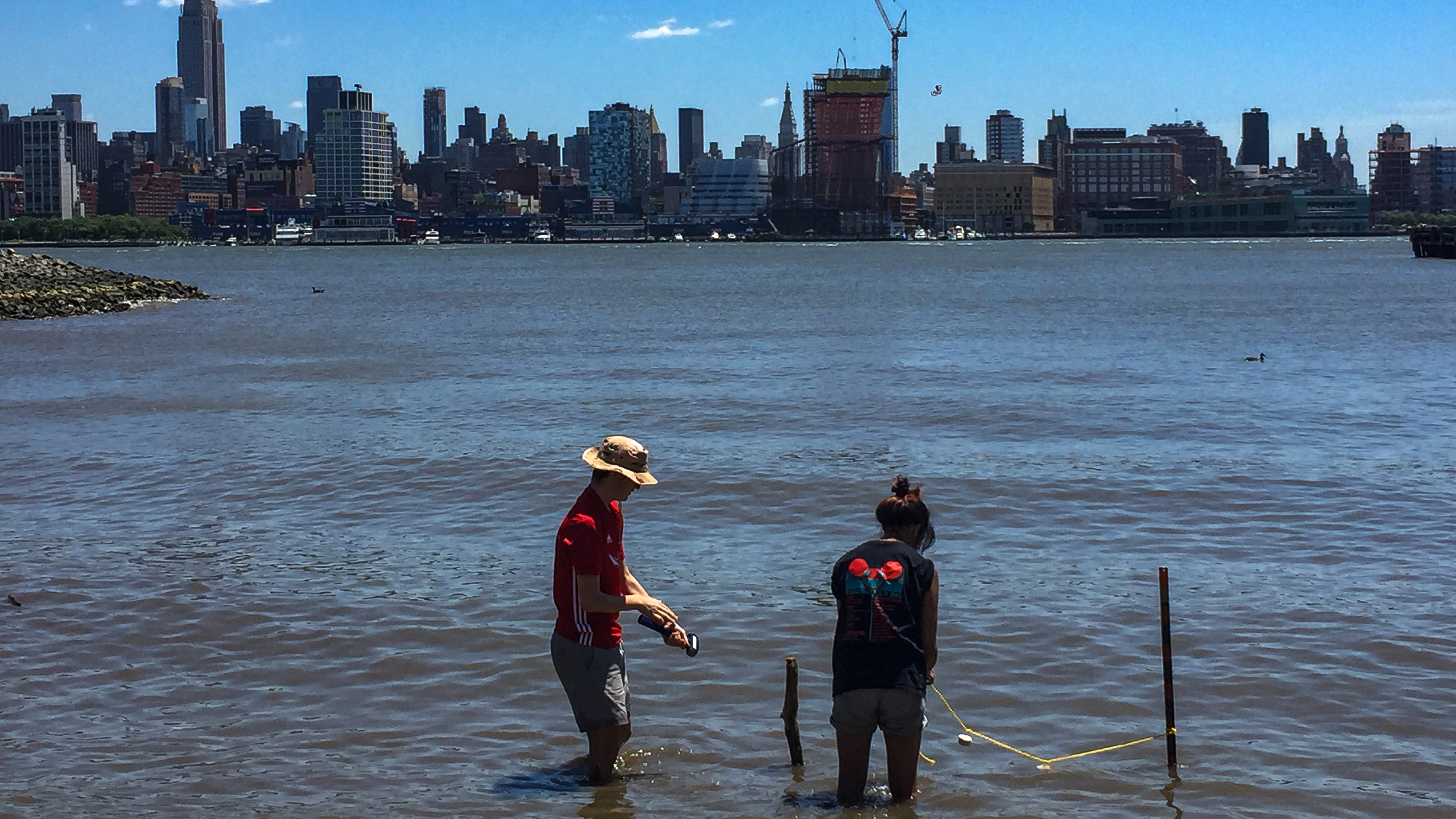 Two people wade in shallow water on the shore of Hoboken, New Jersey, with the New York City landscape in the background. 