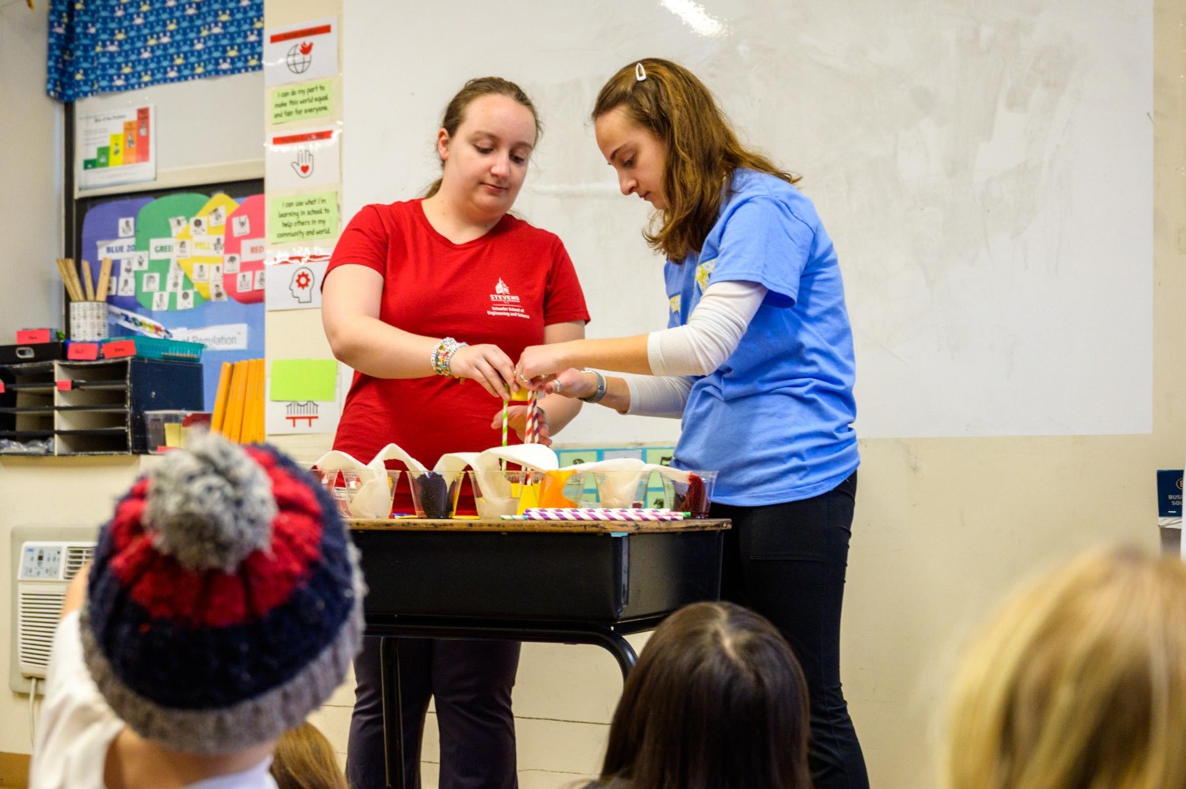 Two people stand at a table conducting a science experiment as children look on.