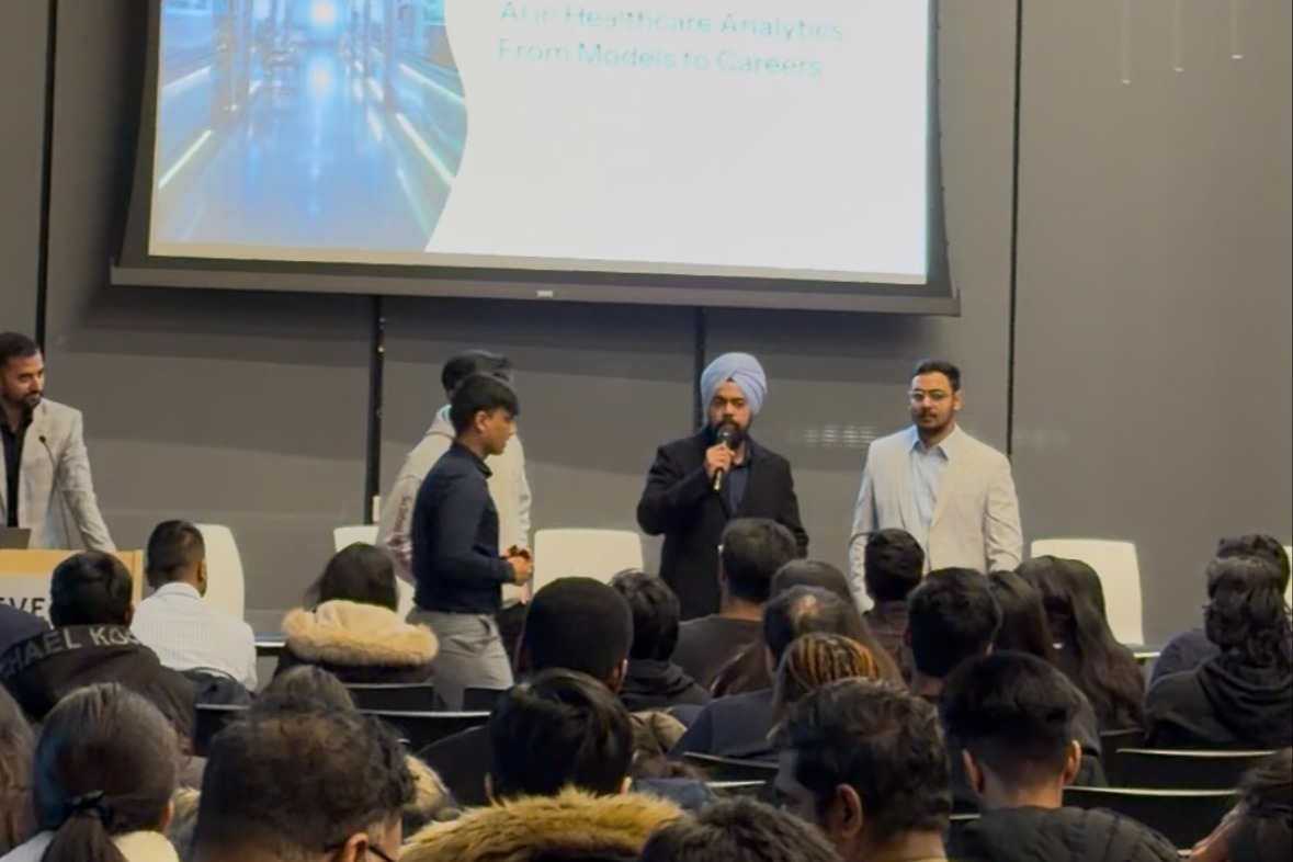 A panel of three speakers presents to an audience in a modern auditorium, with a large screen behind them displaying "AI in Healthcare Analytics From Models to Careers." A speaker in a turban holds a microphone while two others stand beside him.