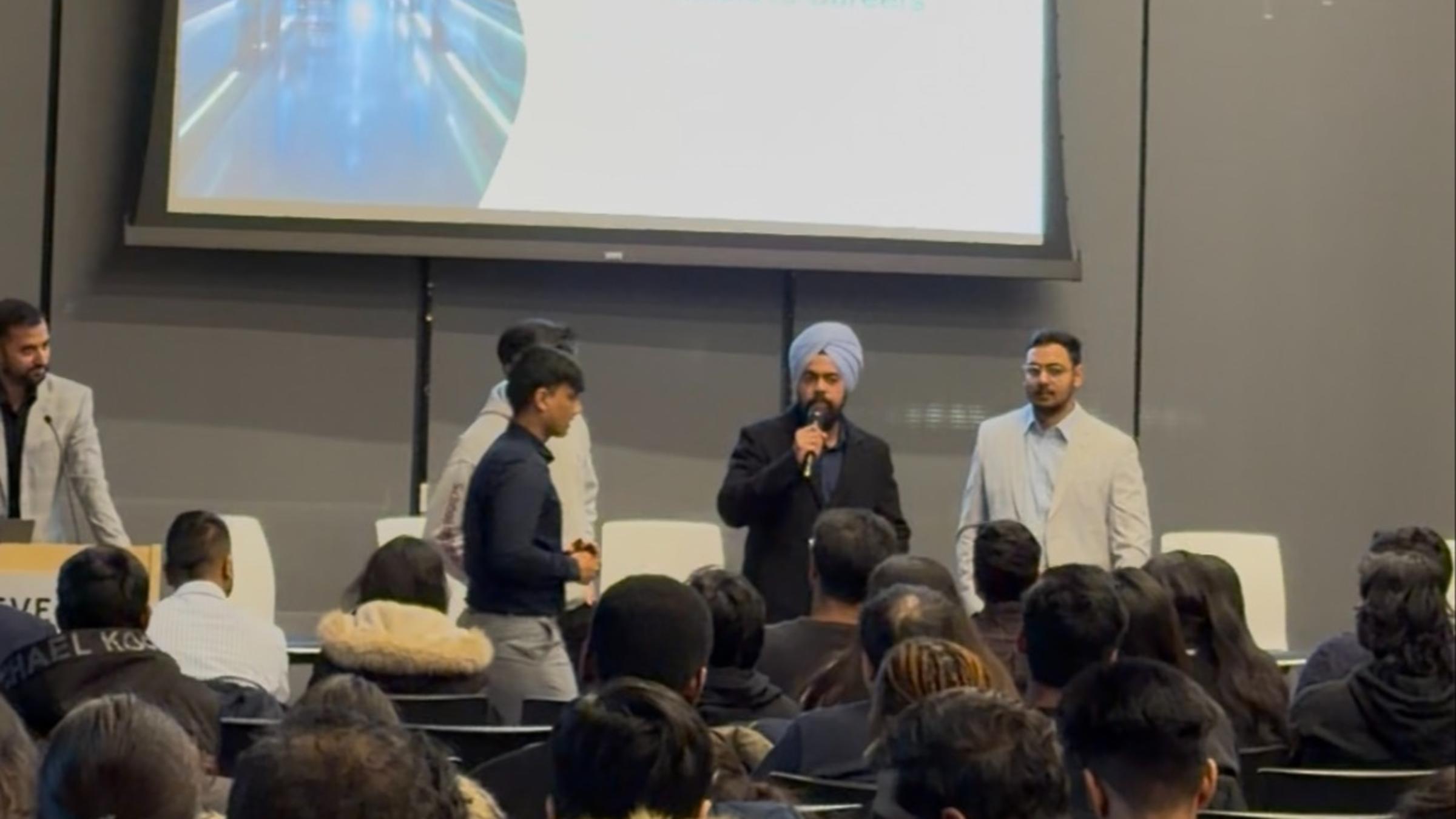 A panel of three speakers presents to an audience in a modern auditorium, with a large screen behind them displaying "AI in Healthcare Analytics From Models to Careers." A speaker in a turban holds a microphone while two others stand beside him.