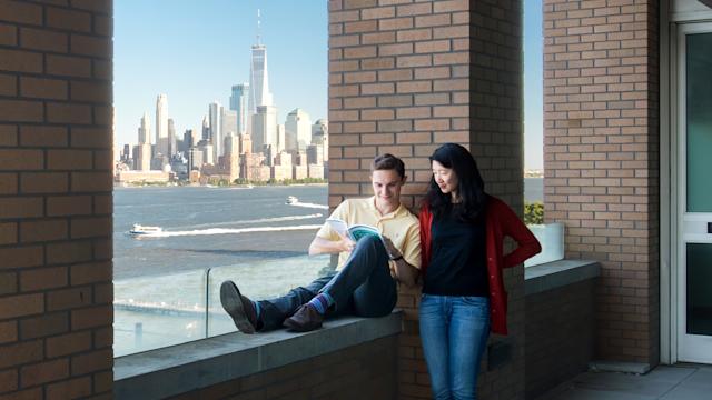 Two students lounge outdoors with the New York city skyline behind them.