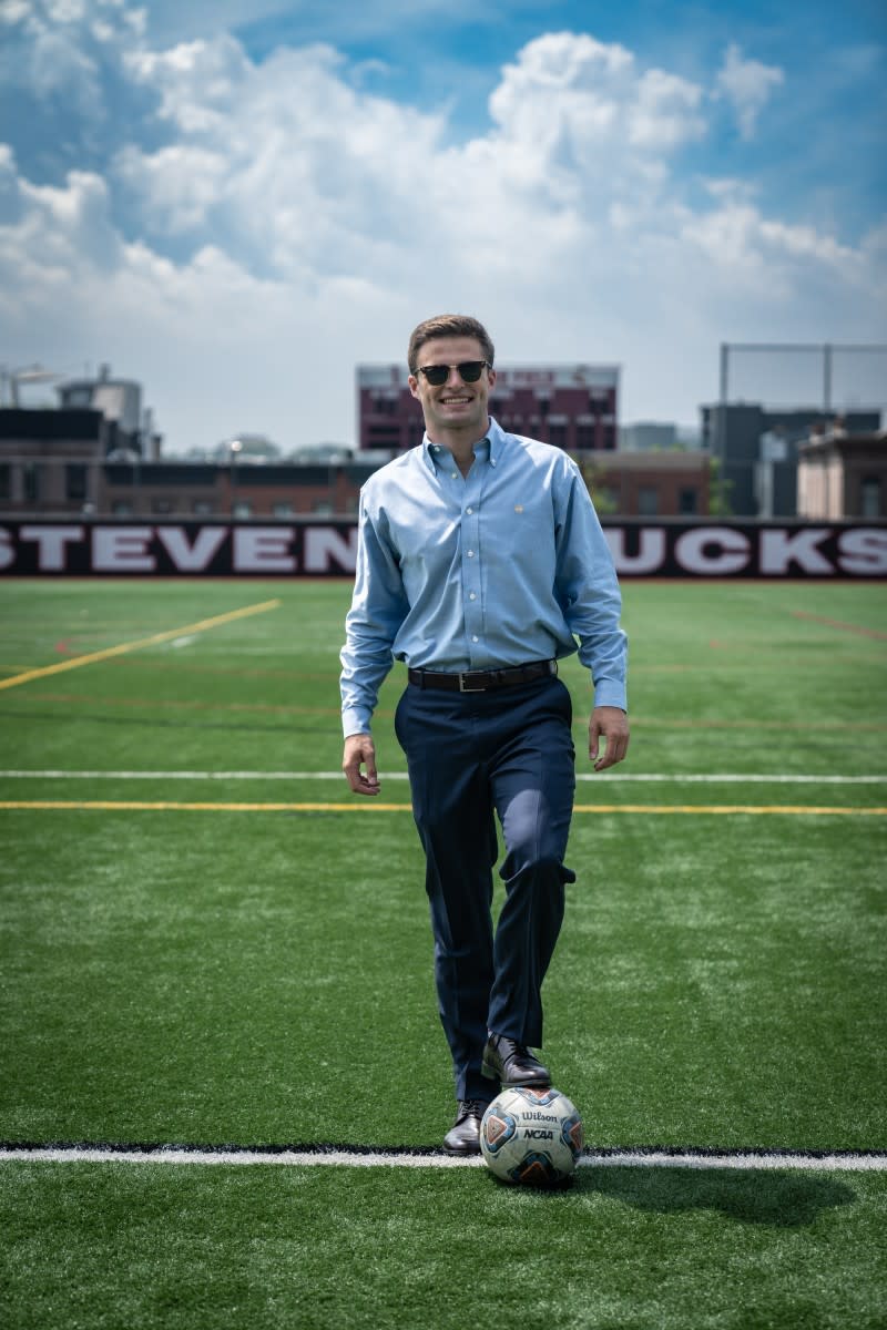 Jay Poskitt is standing at a soccer field in professional attire with a soccer ball underneath his foot