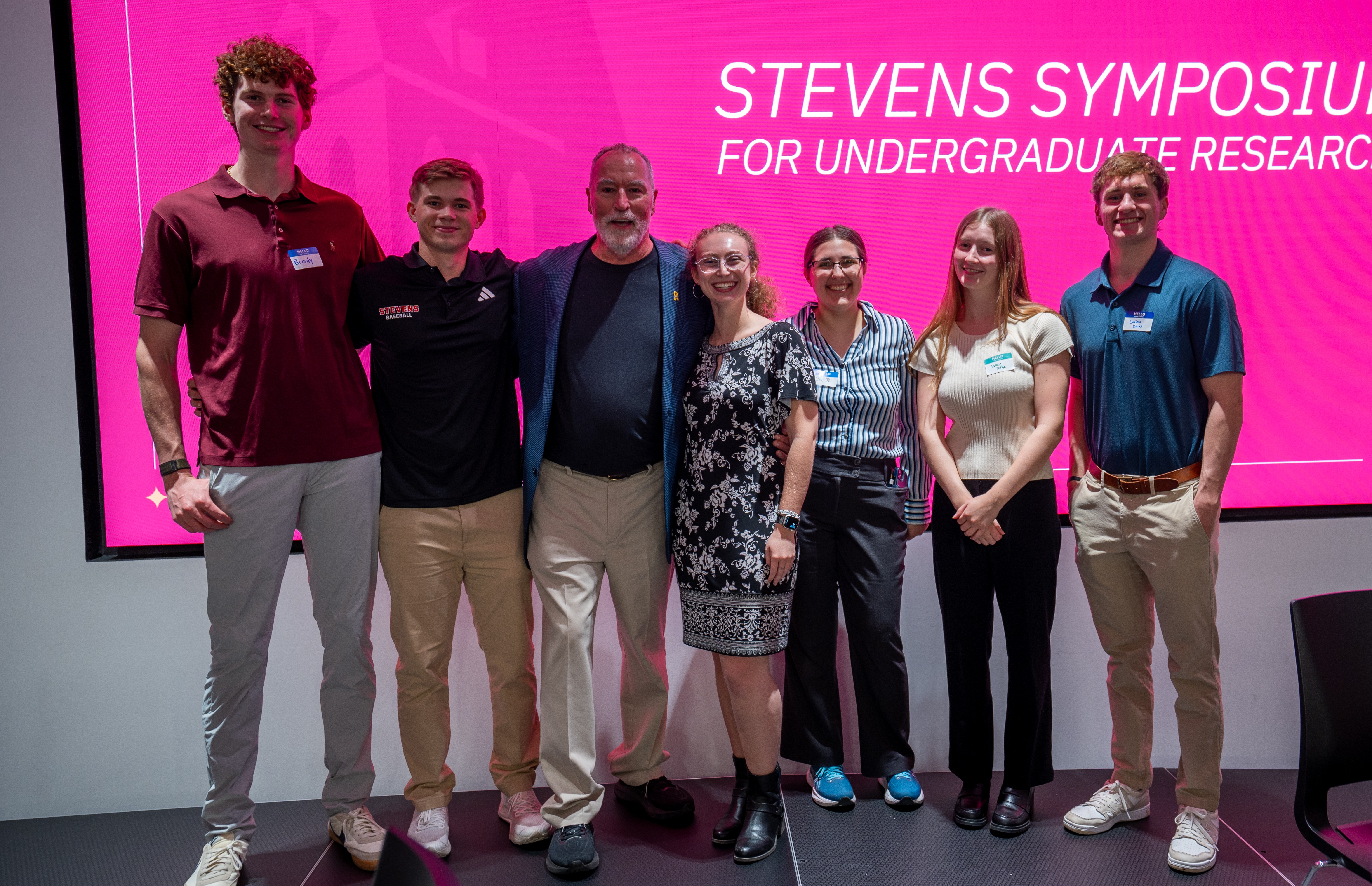 Seven people pose together on stage in front of a large pink screen displaying "Stevens Symposium for Undergraduate Research." The group includes students and faculty in business casual attire, standing in a line and smiling at the camera.