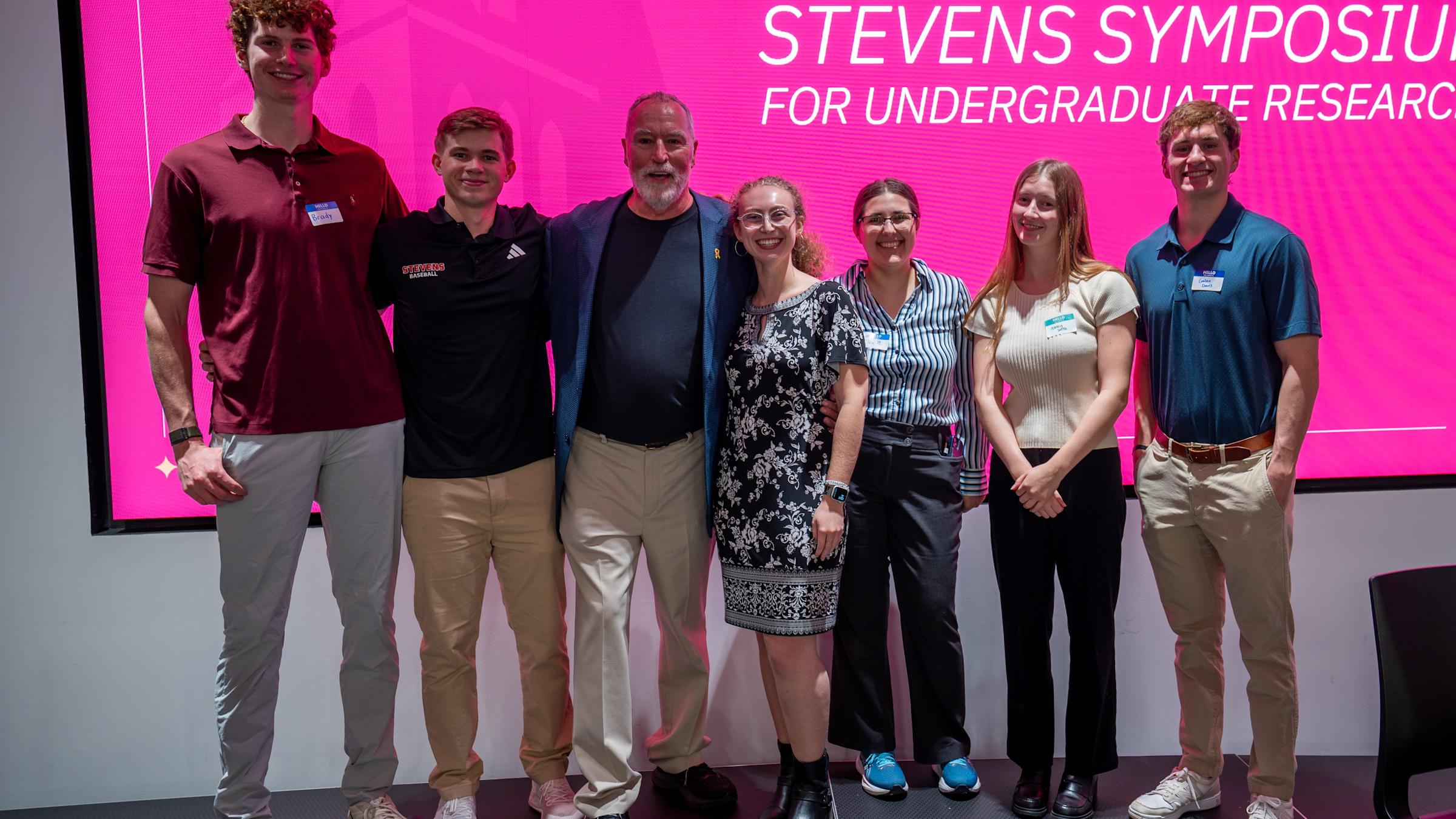 Seven people pose together on stage in front of a large pink screen displaying "Stevens Symposium for Undergraduate Research." The group includes students and faculty in business casual attire, standing in a line and smiling at the camera.