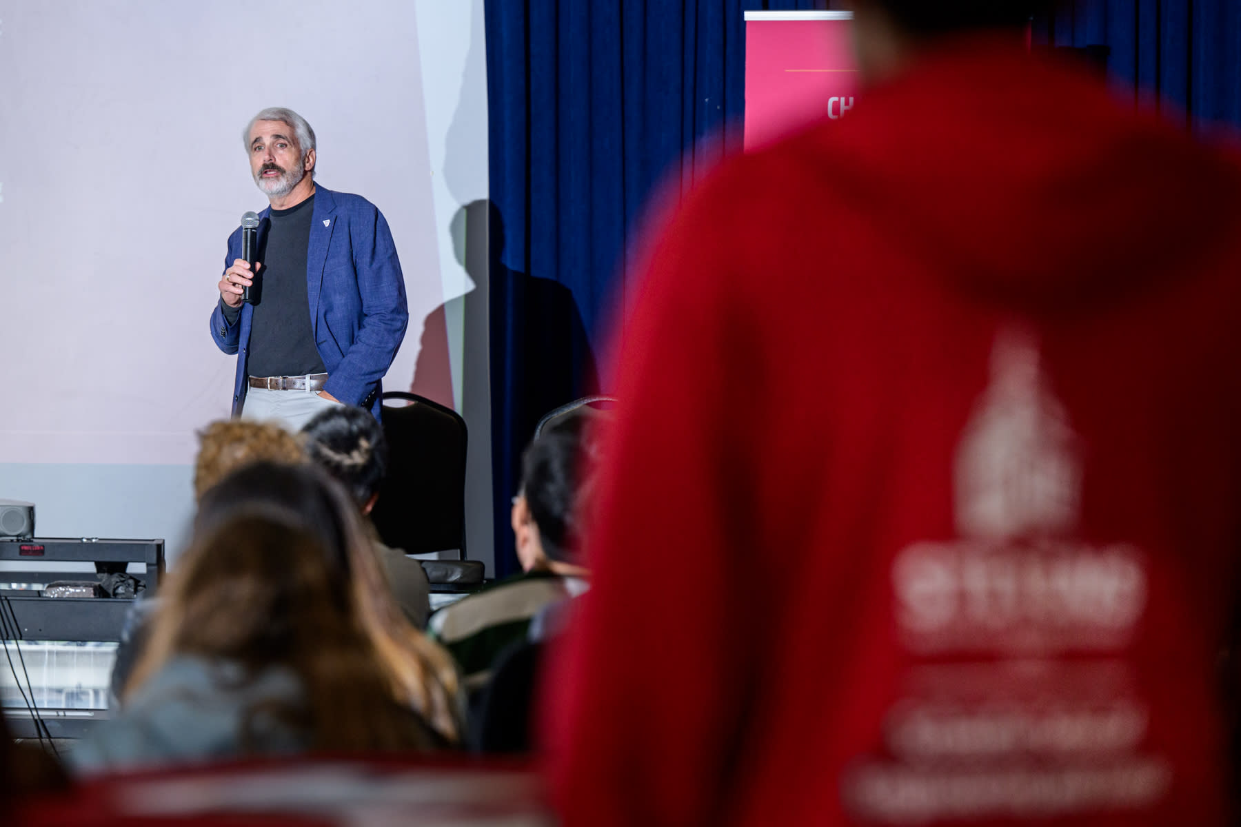 Speaker Kevin Loughran on stage with a microphone while a student wearing a red Stevens hoodie is back-facing in the foreground.