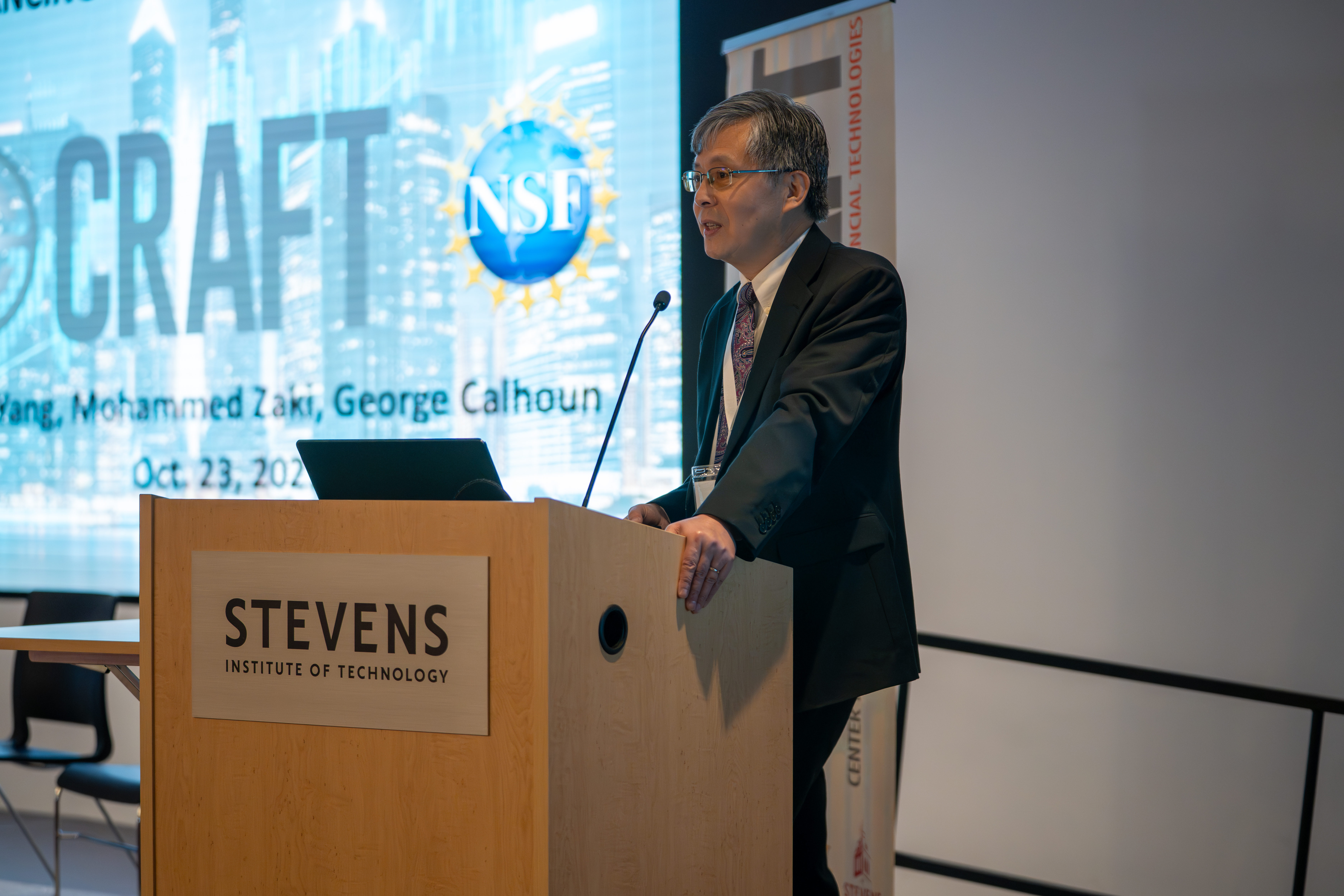 A man in a dark suit and patterned tie presents at a wooden podium bearing the Stevens Institute of Technology logo. Behind him is a projection screen displaying an NSF logo and text referencing Mohammed Zaki and George Calhoun, dated Oct. 23, 2025.
