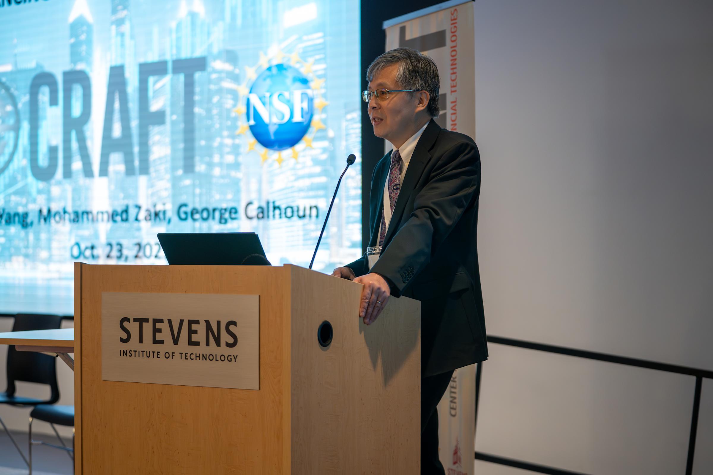 A man in a dark suit and patterned tie presents at a wooden podium bearing the Stevens Institute of Technology logo. Behind him is a projection screen displaying an NSF logo and text referencing Mohammed Zaki and George Calhoun, dated Oct. 23, 2025.