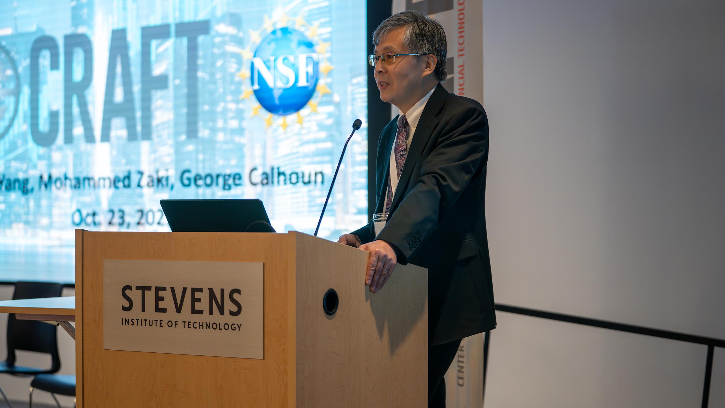 A man in a dark suit and patterned tie presents at a wooden podium bearing the Stevens Institute of Technology logo. Behind him is a projection screen displaying an NSF logo and text referencing Mohammed Zaki and George Calhoun, dated Oct. 23, 2025.