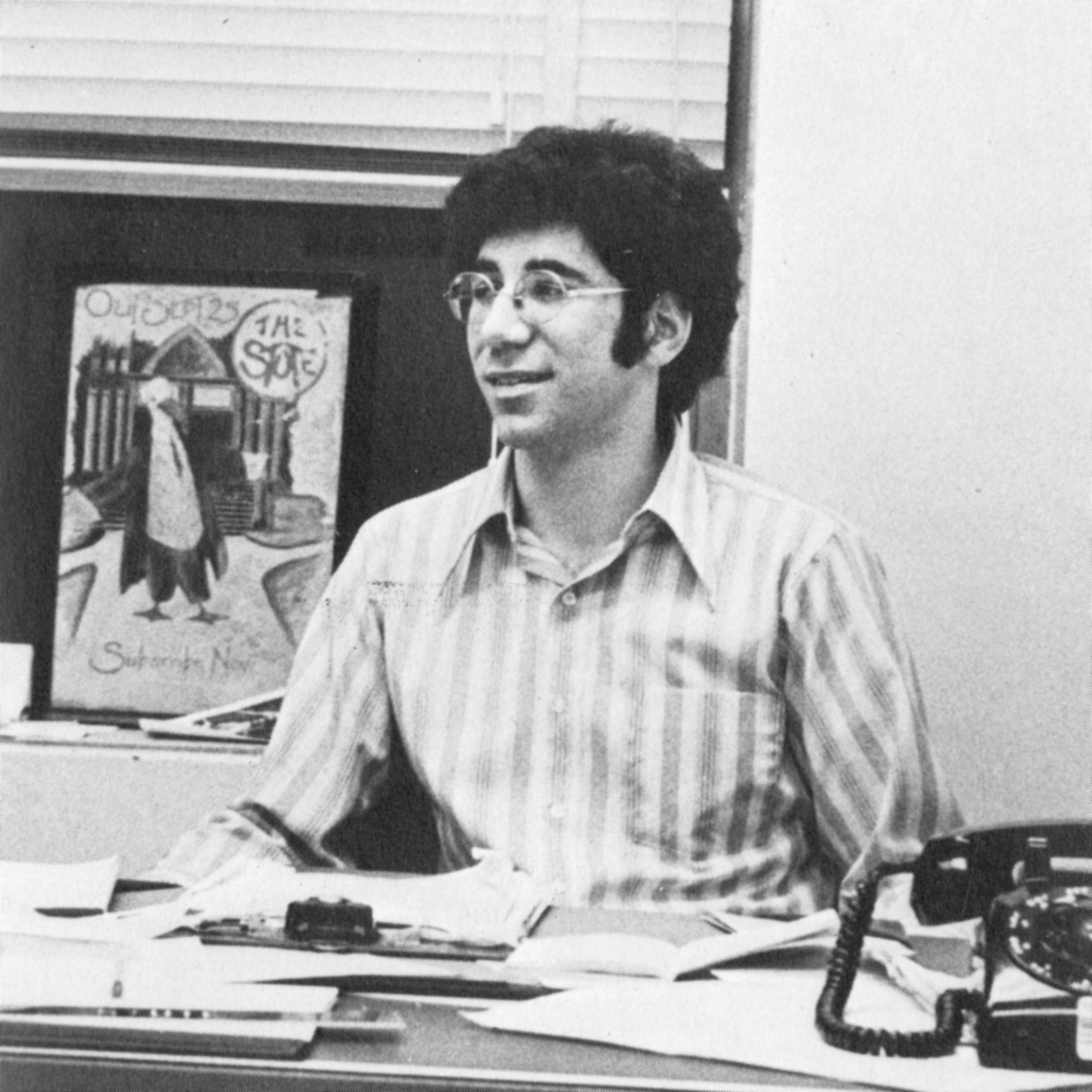 Man in glasses sitting at a desk with papers on it and an old phone.