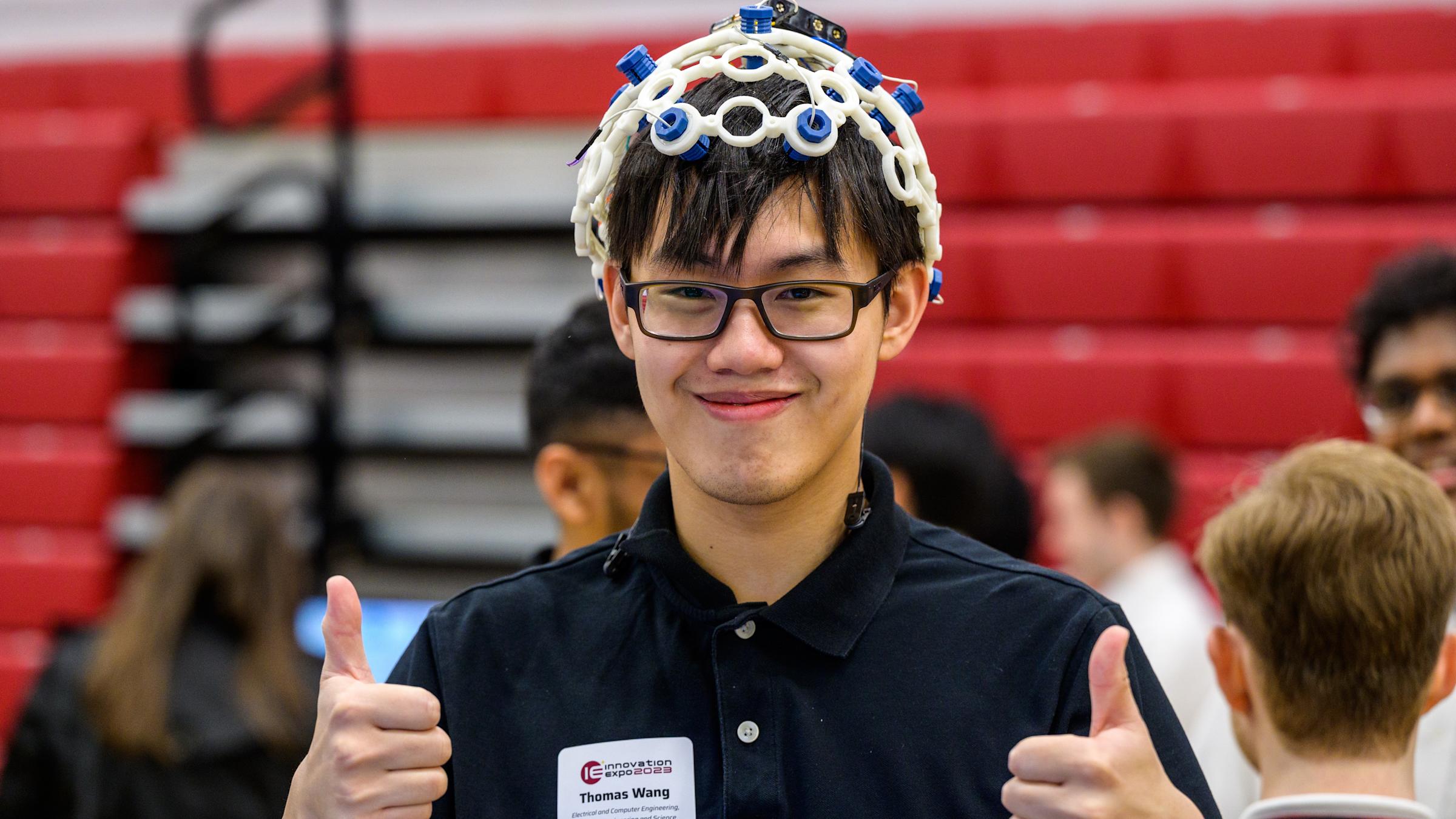 Male student with glasses wearing an experiment on his head with both thumbs up