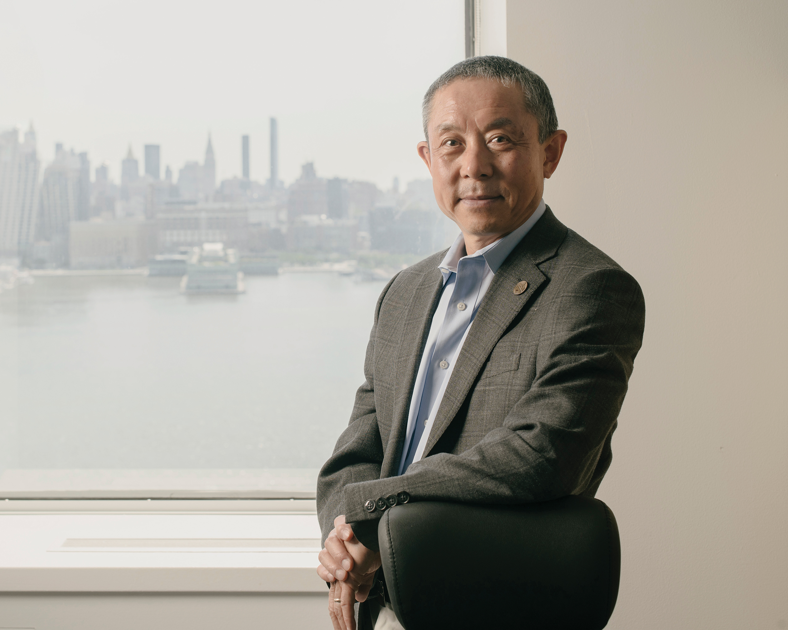 Provost Jianmin Qu poses for the camera in front of a window overlooking the New York skyline.
