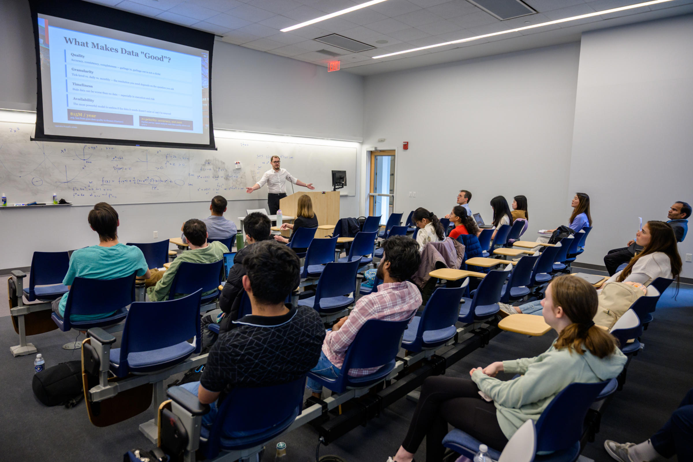 A man standing at a speaking podium talking with a group of students sitting in desk chairs.