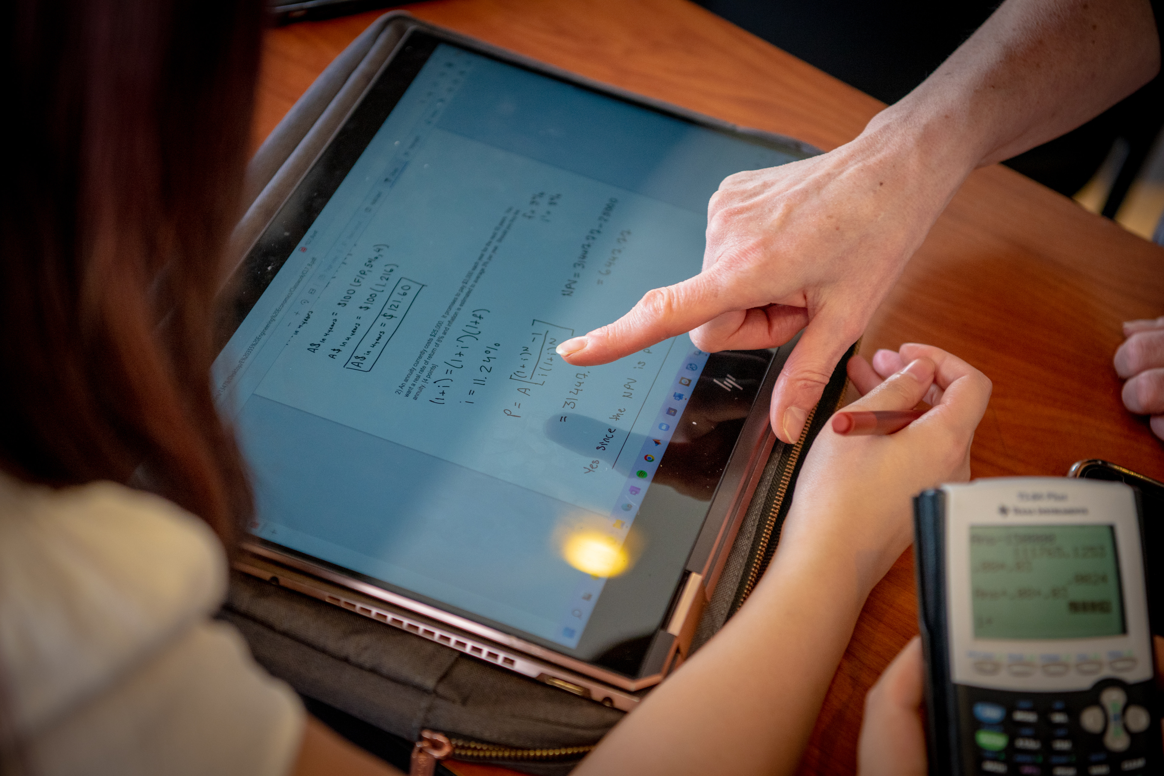 Ipad on a table with math equations with one hand pointing at the screen, another hand on the left and a calculator on the right