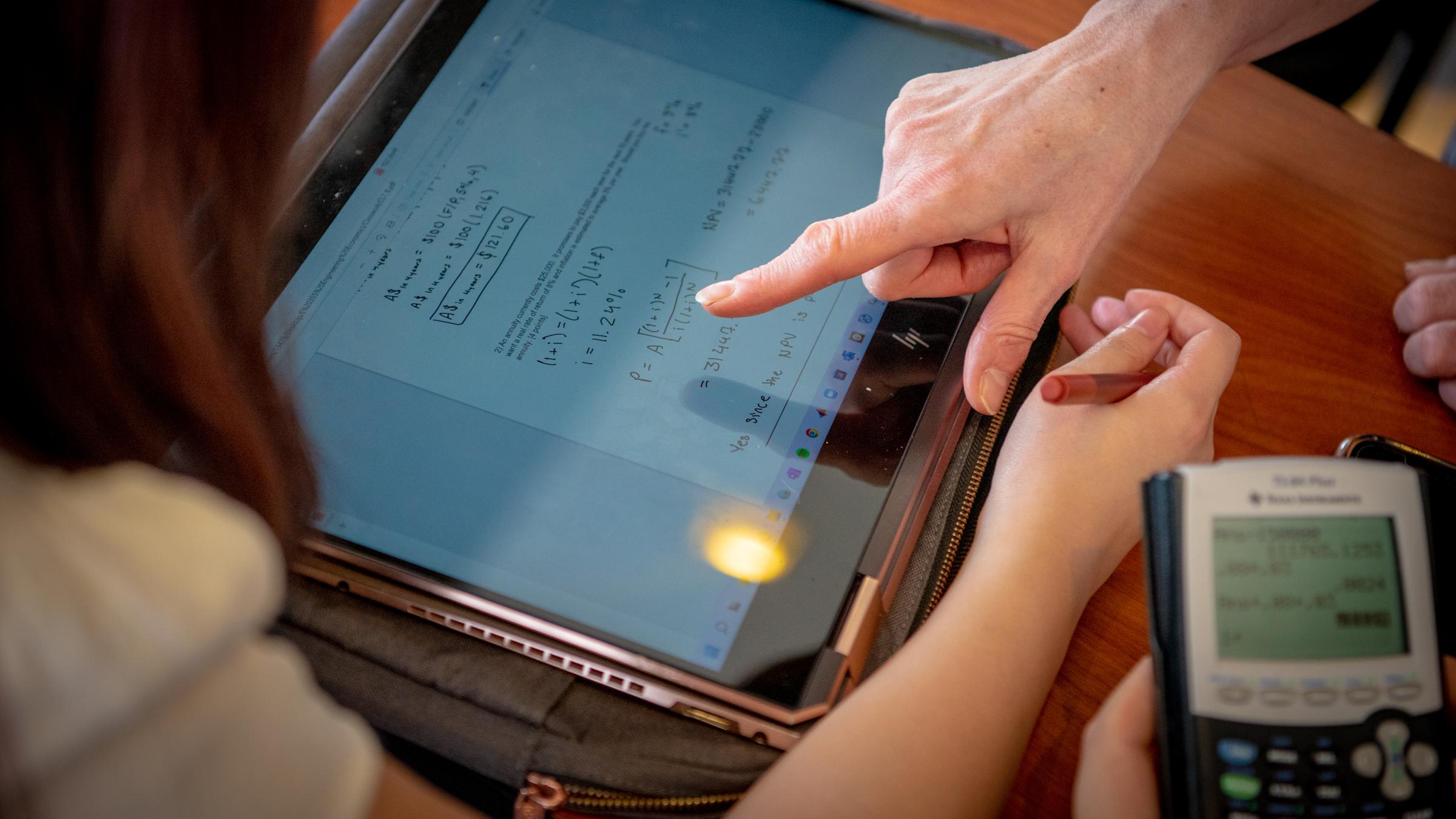 Ipad on a table with math equations with one hand pointing at the screen, another hand on the left and a calculator on the right